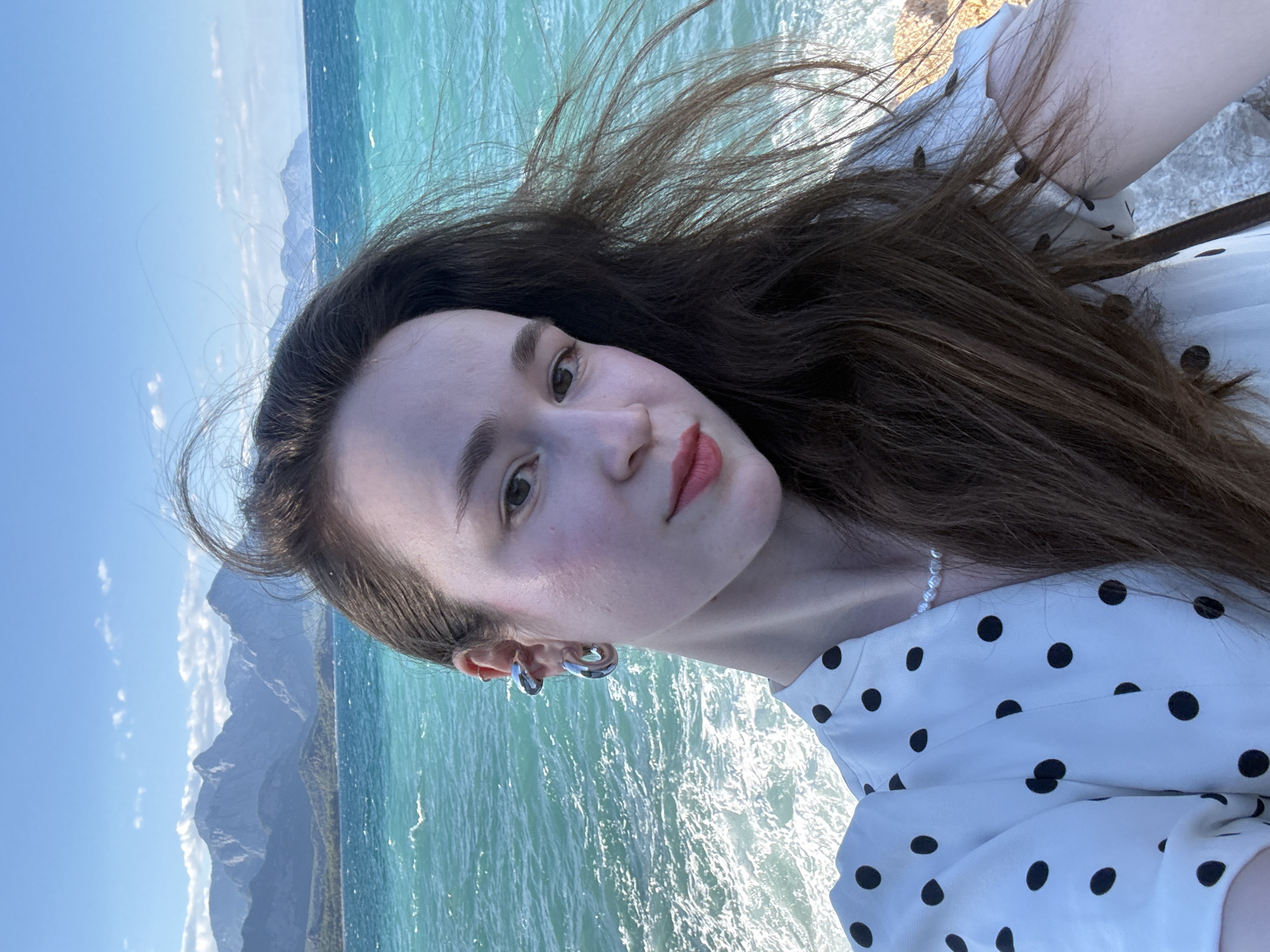 A young woman with long, brown hair stands facing the camera in a selfie. She is wearing a white dress with black polka dots and a pearl necklace. She has on large hoop earrings and ear cuffs. Behind her, the turquoise sea crashes against a rocky shore, with mountains visible in the distance under a bright blue sky. The image appears to be taken in Kemer, Turkey, known for its beautiful coastline and mountainous backdrop.