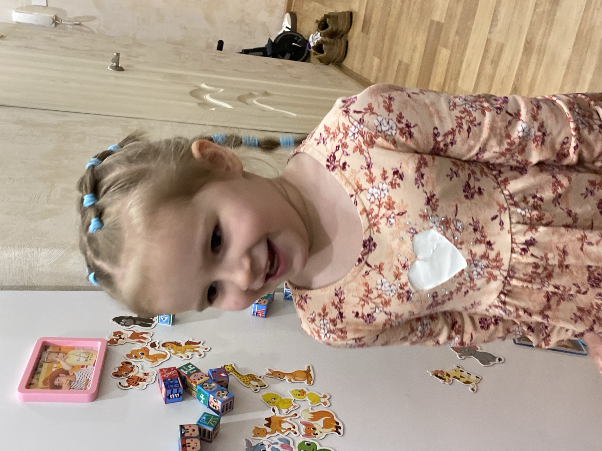 A young girl is playing with toys on a white surface. The girl has blonde hair styled with blue hair ties. She is wearing a floral-patterned dress with a white heart-shaped patch on it. The toys include a pink tray with images inside, animal stickers, and alphabet blocks. The background includes a wall and a wooden floor. The location is likely in Saint Petersburg, Russia, indicated by the Cyrillic letters on some of the alphabet blocks.