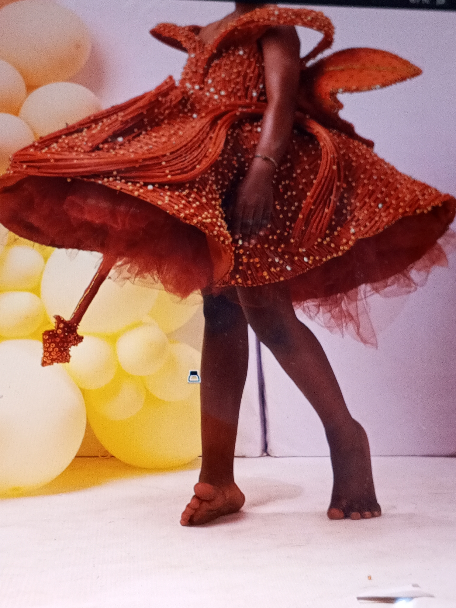 A person with dark skin is wearing an elaborate red dress embellished with small beads. The dress has a ruffled bodice and a full, layered skirt with a tulle underlay. The person's feet are bare, with the right foot turned inward. The background consists of a white floor and a wall behind. To the left of the person is a cluster of light yellow balloons. The dress has an extension. The location appears to be a studio setting. The city, Badagry, Nigeria is not readily apparent from the photo.