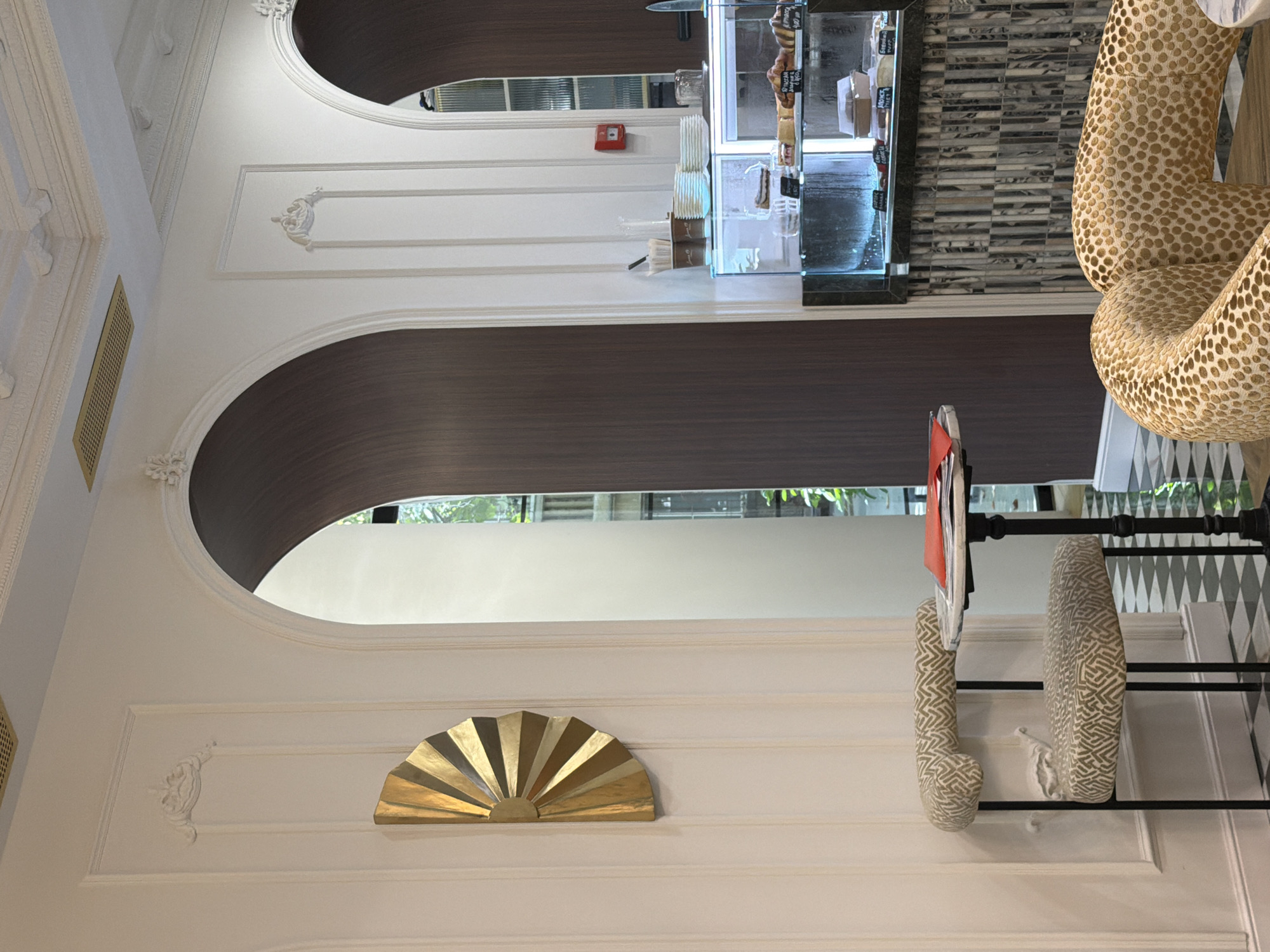 The image depicts the interior of a cafe with a distinctly elegant and classic design, featuring arched doorways and ornate wall molding. On the left, a gold fan-shaped wall decor is mounted on a fluted white wall. In the foreground, a small, round table with a black base holds a stack of papers with a red napkin. Next to it is a patterned armchair with a light-colored fabric featuring a geometric design.
Further into the cafe, a glass display case showcases baked goods. To the left of the display case, there are stacked white plates, two paper cups with lids, and a clear vase. A red fire alarm is visible on the wall. The floor has a black and white checkered pattern. In the background, through an arched opening, a glimpse of another seating area and a window with greenery is visible. Another armchair, with a leopard-like print, is partially visible in the bottom right corner. The cafe appears to be located in Kaspiysk, Russia. There are no people visible in the image.