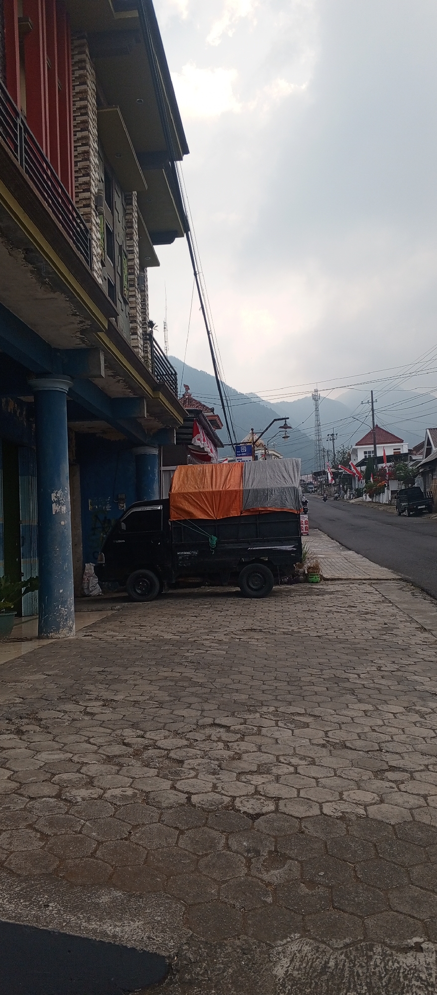 A black pickup truck is parked on a hexagonal cobblestone sidewalk adjacent to a multi-story building with a red facade and blue columns. The truck's bed is covered with an orange and grey tarp, secured with ropes. The building appears to be a commercial establishment with windows on the upper floors. A street runs alongside the sidewalk, lined with various buildings and streetlights. In the background, rolling hills and mountains are visible under a cloudy sky. A cell tower stands prominently in the distance. The scene is set in the city of Poncol, Indonesia.