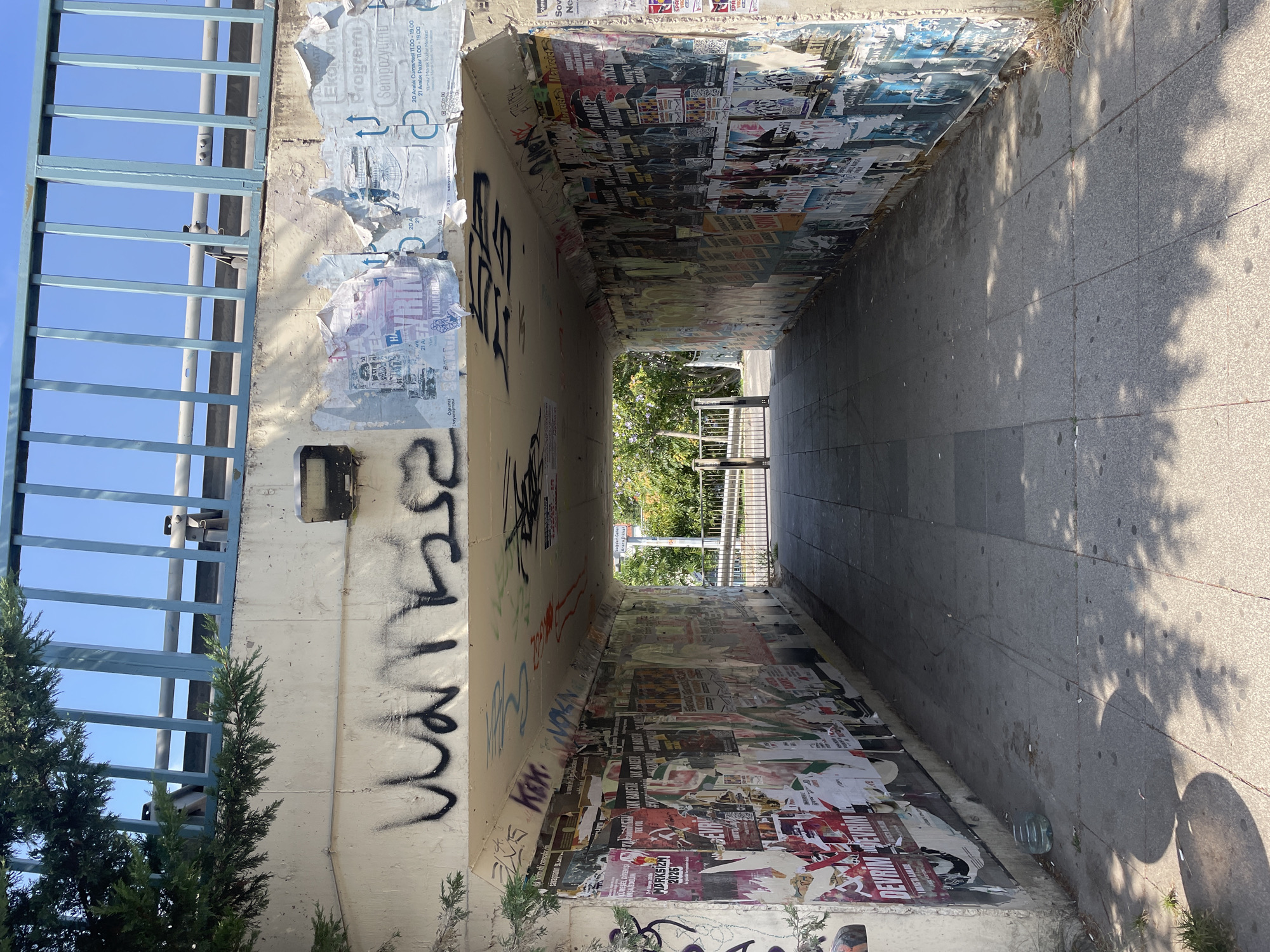 This image depicts a pedestrian underpass in Kadıköy, Turkey, likely located near a busy street or intersection given the presence of a bridge overhead. The underpass walls are extensively covered in a collage of posters, many of which are torn and peeling, suggesting a high volume of public advertising and event promotion. The floor of the underpass is paved with gray concrete tiles. Graffiti is visible on the concrete ceiling and upper walls, with some of it appearing to be spray paint.
On the left side of the underpass, a portion of a tree with green foliage is visible, partially obscuring the top left corner of the structure. The bridge above has a blue railing and is supported by a concrete structure. Sunlight filters into the underpass, casting shadows on the ground. The exit of the underpass is visible in the distance, leading to an outdoor area with more greenery and a metal fence. There are no people present in the underpass. The overall impression is one of urban decay and active public space.