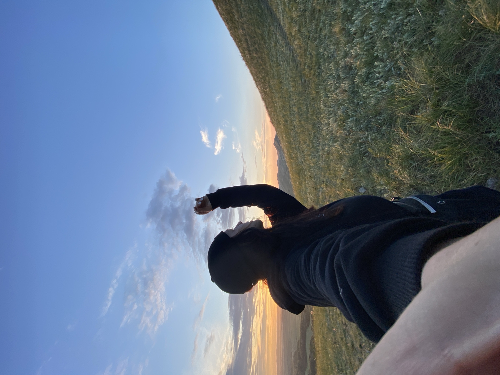 A person stands on a grassy hill, looking towards a sunset over a valley. The person is wearing a black beanie and a black hoodie, and their right arm is raised. The sky is a blend of blue and orange, with scattered clouds. The hill is covered in green grass and some low-lying vegetation. In the distance, mountains are visible. The overall scene is serene and natural. The location is specified as Абзаново, Russia.