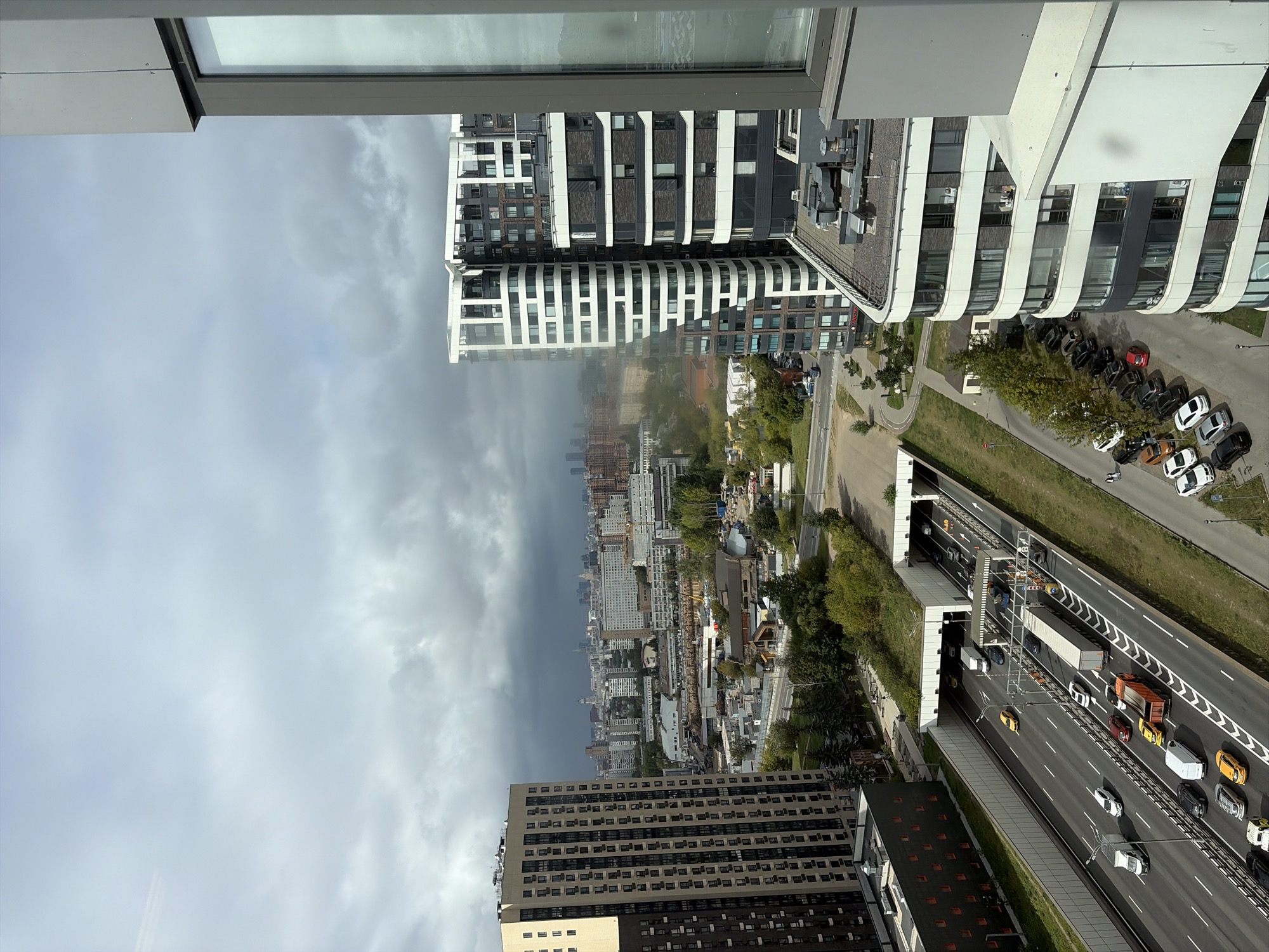 The image shows a view of the Moscow region from a height, likely from an apartment or office building. In the foreground, a modern residential building with a curved facade and white and dark panels is visible on the right. To the left, a tall, rectangular apartment building with beige and dark panels dominates the left side of the frame.
A multi-lane highway runs through the middle ground, with cars, trucks, and a bus visible. There is a tunnel entrance on the highway, covered with a green roof, and a toll plaza. Further in the distance, a sprawling urban landscape of various buildings, including some high-rise structures, stretches to the horizon. The sky is overcast with dark, dramatic clouds. The scene appears to be a residential and commercial area within the city of Moscow, Russia, characterized by contemporary architecture and significant infrastructure.