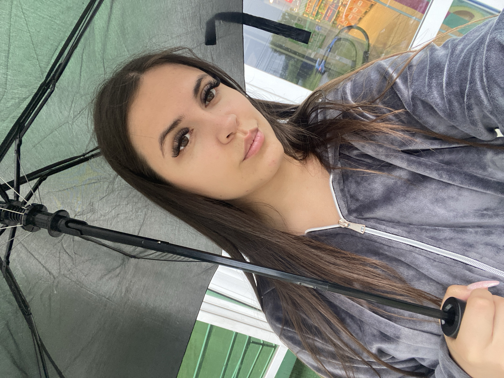 A young woman with long, dark brown hair and long, dark eyelashes is holding an open black umbrella. She is wearing a gray velour zip-up hoodie. She is holding the umbrella in her right hand, with her left hand visible near the bottom of the umbrella shaft. The background appears to be a storefront with a large window reflecting a street scene. The woman is looking towards the camera. The image was taken outdoors, likely in a city, as suggested by the reflections in the window which show store aisles and possibly streetlights. The location is Chaikovsky, Russia, indicated by the file name which contains "Chaikovsky".