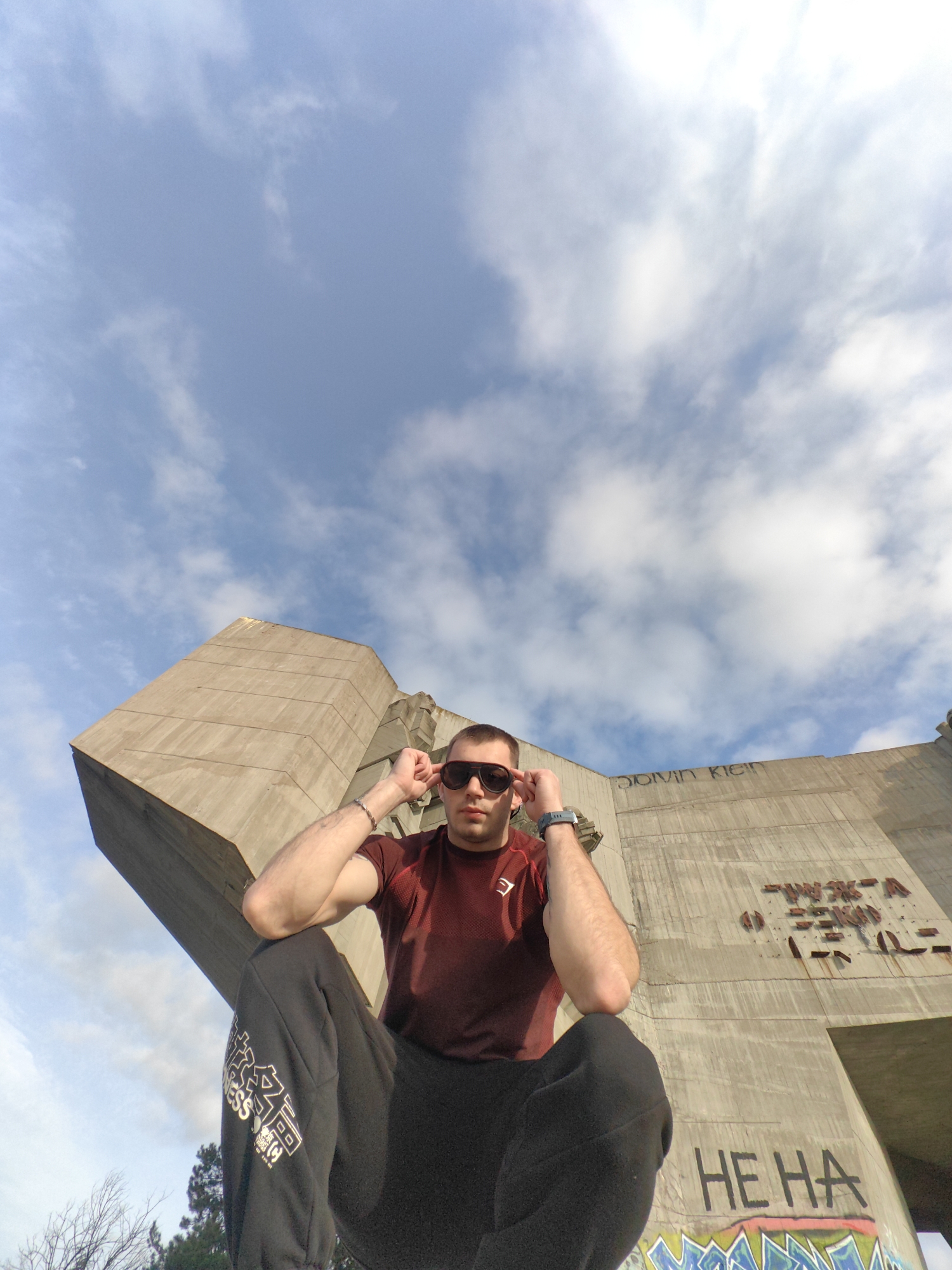 A low-angle, medium shot depicts a man with short, dark hair and a maroon shirt, wearing sunglasses. He is sitting with his legs folded, his arms raised to adjust his sunglasses. His wrists are adorned with a bracelet on the left and a watch on the right. He's wearing black sweatpants with white lettering on the left thigh.
Behind him, a large concrete structure dominates the background, marked with graffiti, including names "DOMIN KLEIN" and phrases "HE HA." Above the structure, the sky is partly cloudy. The location is the Monument to the Bulgarian-Soviet Friendship in Varna, Bulgaria.