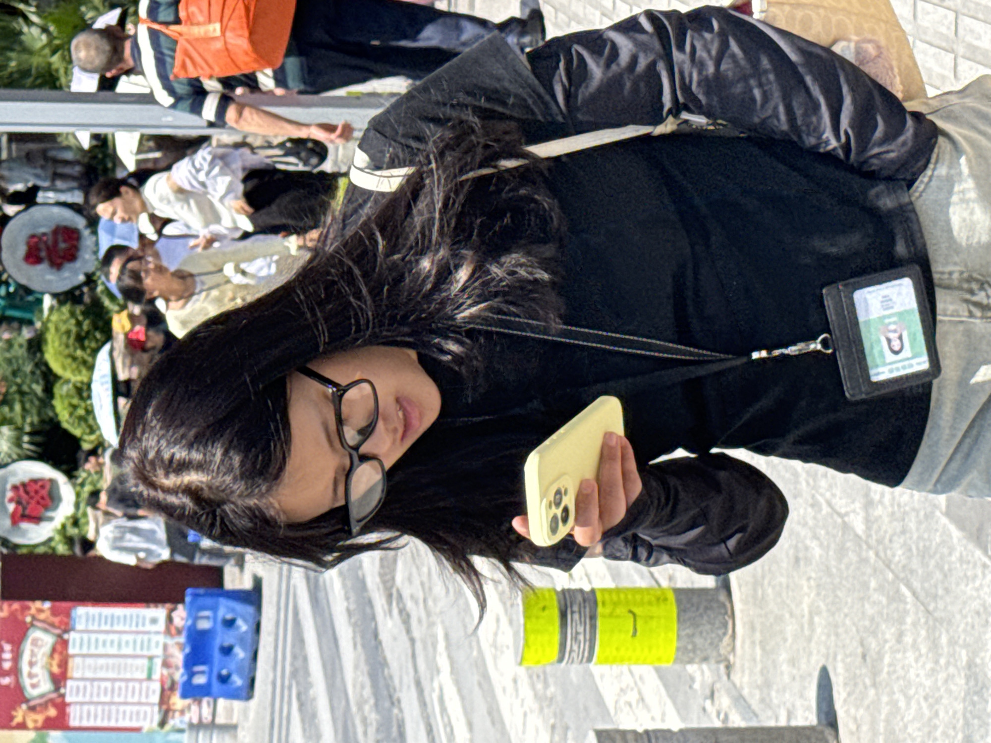 A person is looking at a yellow mobile phone, with a black jacket. The person is wearing glasses. There is a lanyard hanging around the person's neck. A black wallet/card holder is attached to the lanyard. The setting is outdoors. Other people are in the background, and there is a red sign. The image was taken in Shapingba District, China.