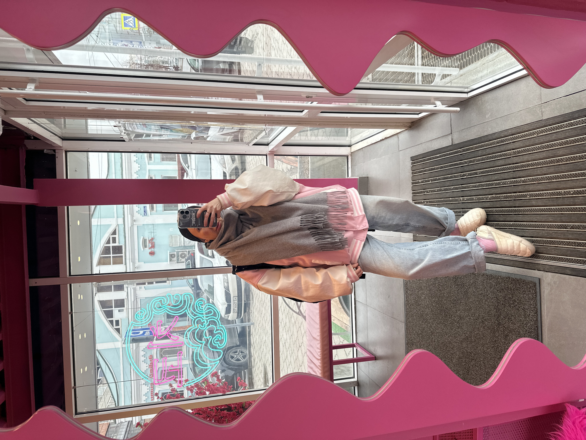 The image captures a mirror selfie taken by a person inside a brightly lit establishment in Perm, Russia. The subject, seen from the waist down to the head, is dressed in a grey hooded top, a grey fringed scarf, a light pink shirt or hoodie underneath, light blue baggy jeans, light pink socks, and cream-colored slip-on shoes. A cream-colored bag is visible over one shoulder. They hold a black smartphone, with a case displaying text like "XOOXIA," to take the photo, reflecting their face.
The interior features distinctive wavy pink decorative elements overhead and along a side wall, with a pink bench also present. The floor is covered with grey tiles and dark grey textured mats. Through the large glass facade, an urban street scene is visible. Buildings with light blue and grey exteriors and various windows are seen across the street. Parked cars, including a white car with a partially visible license plate "H313PB," are outside. A striking turquoise and pink neon sign, displaying stylized Chinese characters (possibly related to "ice" or "sweet"), is visible through and reflected in the glass. Red artificial flowers add color to the foreground outside the window. Reflections in the upper glass panes show a blue road sign, likely a pedestrian crossing sign. The scene is illuminated by natural daylight.