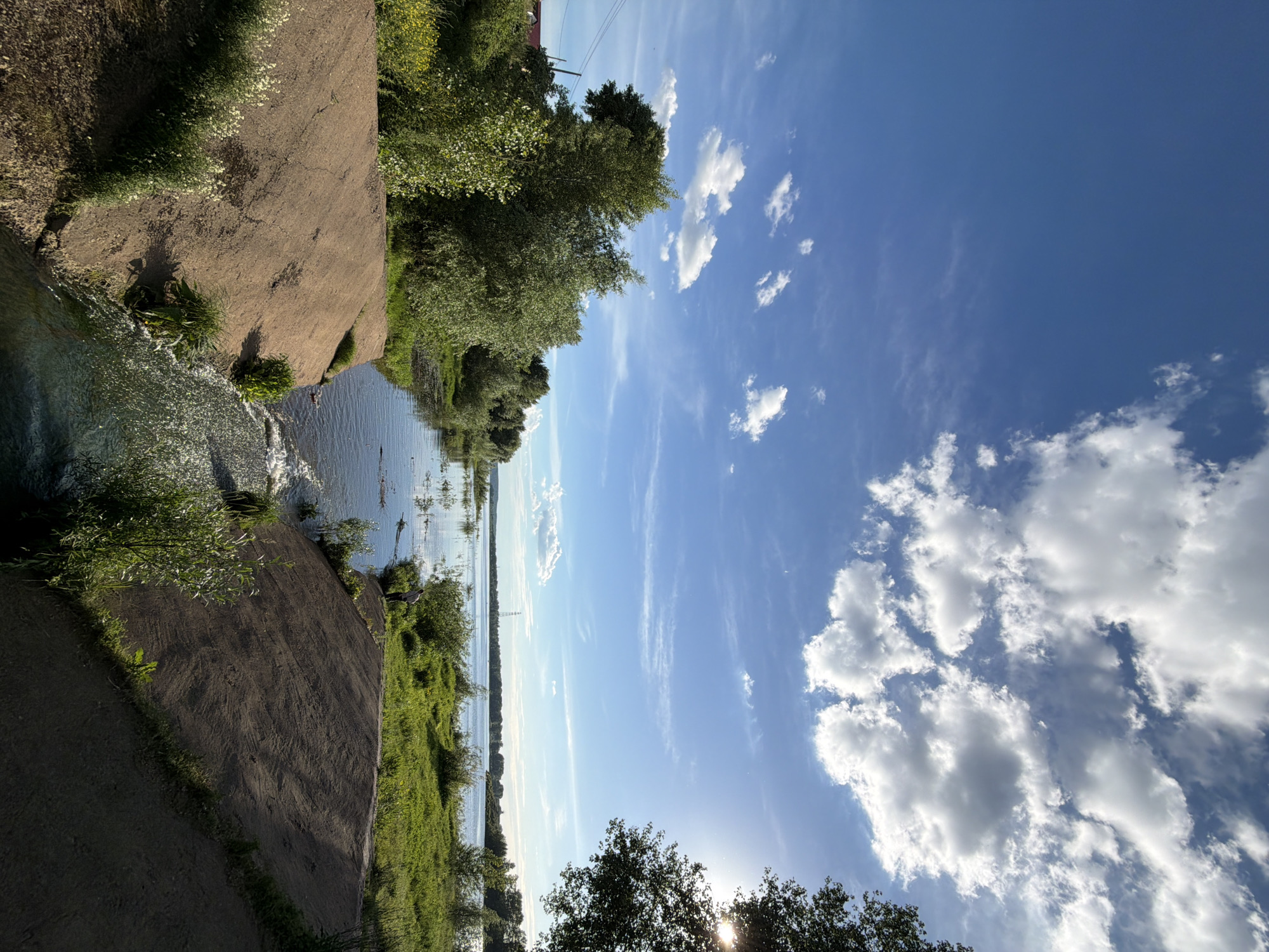 The image captures a scenic view of a waterway and its surroundings on a sunny day, likely in Chaikovsky, Russia. The foreground features a man-made embankment made of dirt, with small plants and white wildflowers growing along its edges. A narrow channel of water cascades down from the embankment, creating a small waterfall. This channel leads into a larger body of water, possibly a river or reservoir, which extends towards the horizon.
The waterway is flanked by lush green vegetation, with trees and bushes lining its banks. In the distance, a treeline is visible, and further still, the horizon is marked by a faint line of buildings or structures. The sky is a vibrant blue, dotted with fluffy white clouds, indicating fair weather. The sun is bright, casting a gentle light and creating a slightly hazy effect in some parts of the image.
A person is visible standing near the edge of the larger body of water, facing away from the camera, seemingly observing the landscape. The overall impression is one of tranquility and natural beauty, with a touch of human intervention in the form of the embankment and channel. The environment appears to be a natural or semi-natural park area, popular for outdoor recreation.