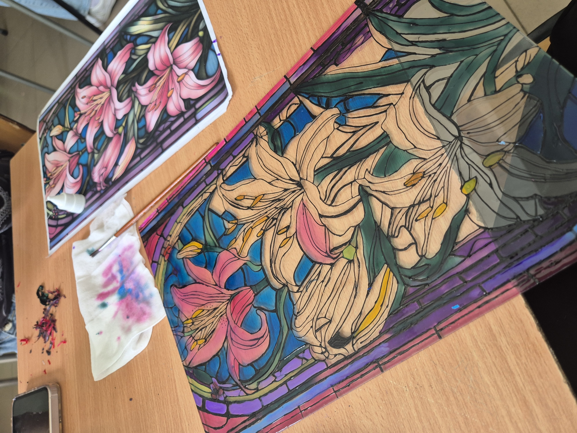 The image displays an indoor art workspace in Pavlikeni, Bulgaria, featuring a vibrant stained-glass-style artwork in progress. The scene is set on a light wooden table, where artistic creation is clearly underway.
The central subject is a large rectangular piece, likely made of glass or clear acrylic, on which a design of lilies in a stained-glass mosaic style is being painted. The design features multiple lily blooms with sweeping petals and long stamens. Parts of the artwork are vibrantly colored in shades of pink, cream, yellow, and green for the flowers and foliage, set against a background of blues and teals, and framed by a rich purple and magenta border. Other sections of the lilies and background are still uncolored, showing only the dark outlines, or are partially filled, indicating the work is incomplete.
To the upper left, a smaller printed reference image of the same lily design, fully colored and detailed, lies on the table, serving as a guide for the artist.
Surrounding the main artwork are various tools and materials. A small white bottle, possibly containing paint or medium, is near the reference print. A paintbrush rests on a crumpled white cloth that is heavily stained with colorful splotches of pink, blue, and teal paint, suggesting recent use. Nearby, a mobile phone is partially visible, and dried, colorful paint splatters are scattered on the wooden surface, further indicating active creative work. The lighting appears bright and even, illuminating the artistic process. No discernible text is visible within the image.