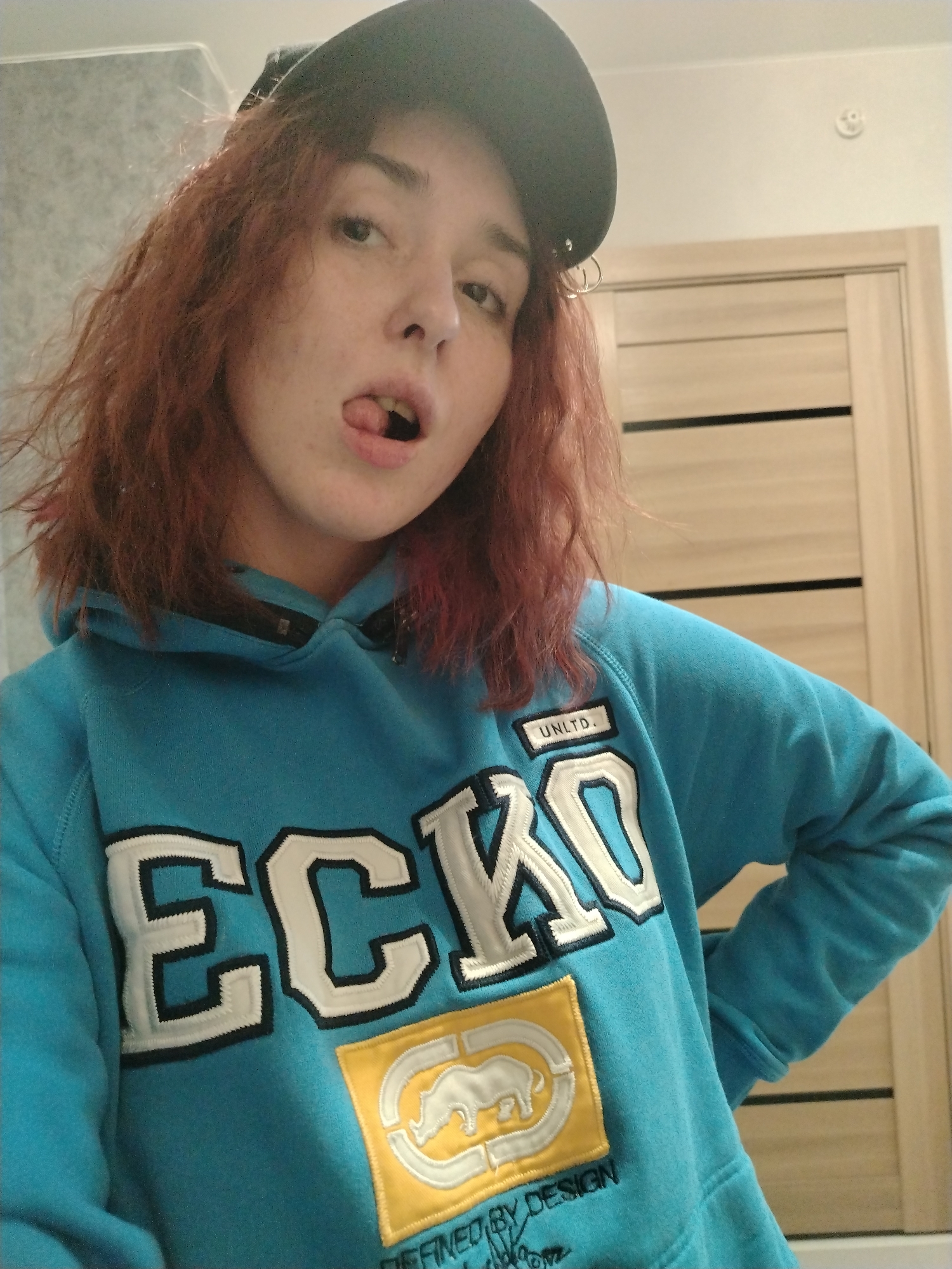 An indoor scene features a single female individual from the upper torso upwards. She has reddish-brown, wavy hair and fair skin with freckles. A dark-colored baseball cap is worn on her head, displaying small silver hoop accessories on the left side. Her mouth is open with her tongue partially extended. She is wearing a light blue hooded sweatshirt adorned with large white and black embroidered "ECKO" lettering across the chest. Below this, a yellow rectangular patch displays a white rhinoceros logo and the text "UNLTD." and "DEFINED BY DESIGN." A small black patch with "UNLTD." is visible near the collar. The background includes a light grey textured wall on the left and a light brown wooden door with two horizontal dark stripes on the right. A white ceiling with a white circular smoke detector is visible in the upper right. No specific location within Izhevsk, Russia, can be determined from the provided image.