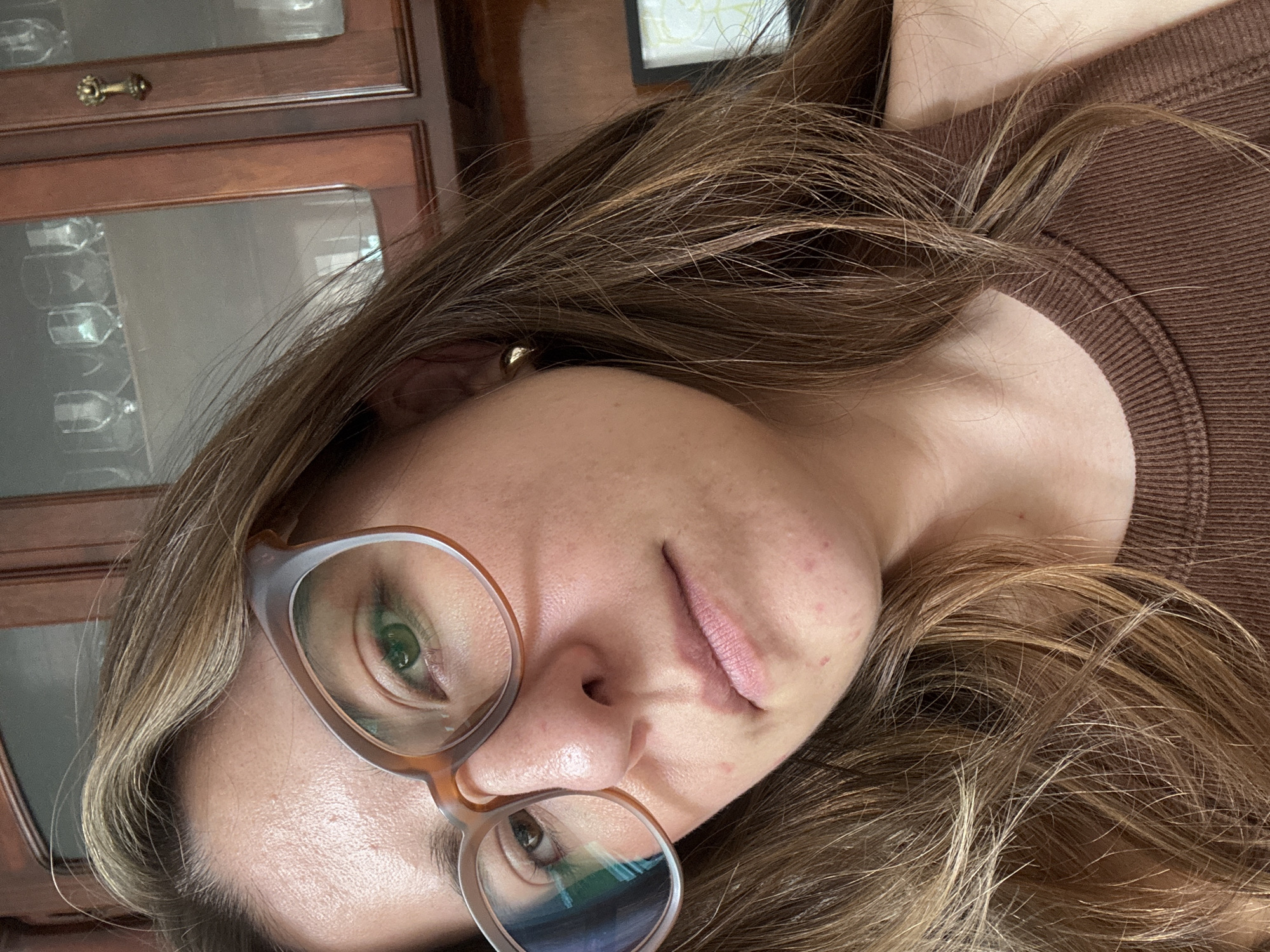 The image presents a close-up portrait of a person, likely female, captured from the chest up. The photograph is oriented sideways, with the subject's head tilted. She has medium-length, wavy brown hair with noticeable lighter highlights, cascading around her face and shoulders. She wears light-framed glasses, through which her green eyes are clearly visible, directed towards the viewer. Her facial expression is neutral, with her lips slightly parted. Her skin shows some minor blemishes on her cheek and chin. A small gold stud earring is visible in her right ear, partially covered by hair. She is dressed in a ribbed, brown top.
The background suggests an indoor, domestic setting. Behind her, a dark wooden cabinet or hutch with glass doors is visible, containing several clear glasses or stemware. A decorative brass drawer pull is seen on the cabinet. In the upper right corner, a portion of a framed picture or artwork with abstract lines can be discerned. Reflections from indoor light sources are visible in her glasses and the cabinet's glass panels. The activity is a direct gaze portrait, possibly a selfie. There is no visible text in the image. This scene was captured in Puebla, Mexico.