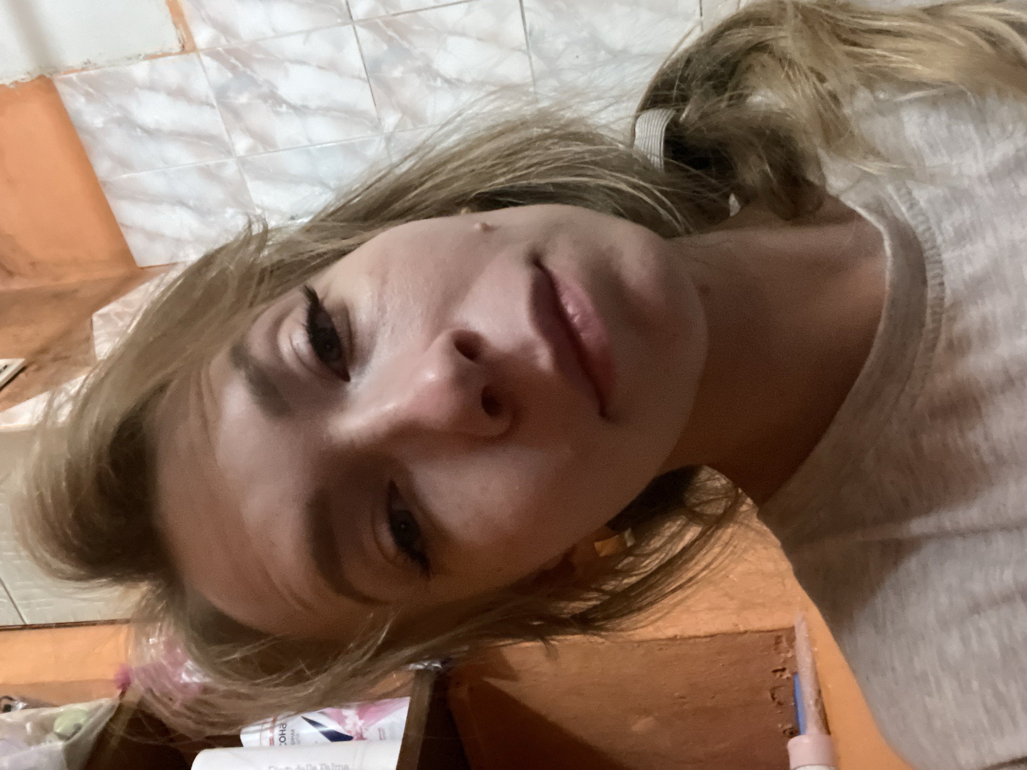 A young, fair-skinned woman with blonde hair pulled back in a ponytail is positioned in the lower half of the frame, close to the camera. She is looking slightly down and to the left. She has eyeliner on her upper eyelids. She is wearing a light-colored shirt. The background consists of light-colored, patterned tiles on the upper left and orange painted walls and a counter area on the lower right. On the counter are several white and pink cosmetic or personal care product containers. The location is likely an indoor bathroom or preparation area, given the tiles and presence of toiletries. There is no verifiable information in the image to confirm the city is Perm, Russia. The image does not offer any data that definitively links it to Perm.