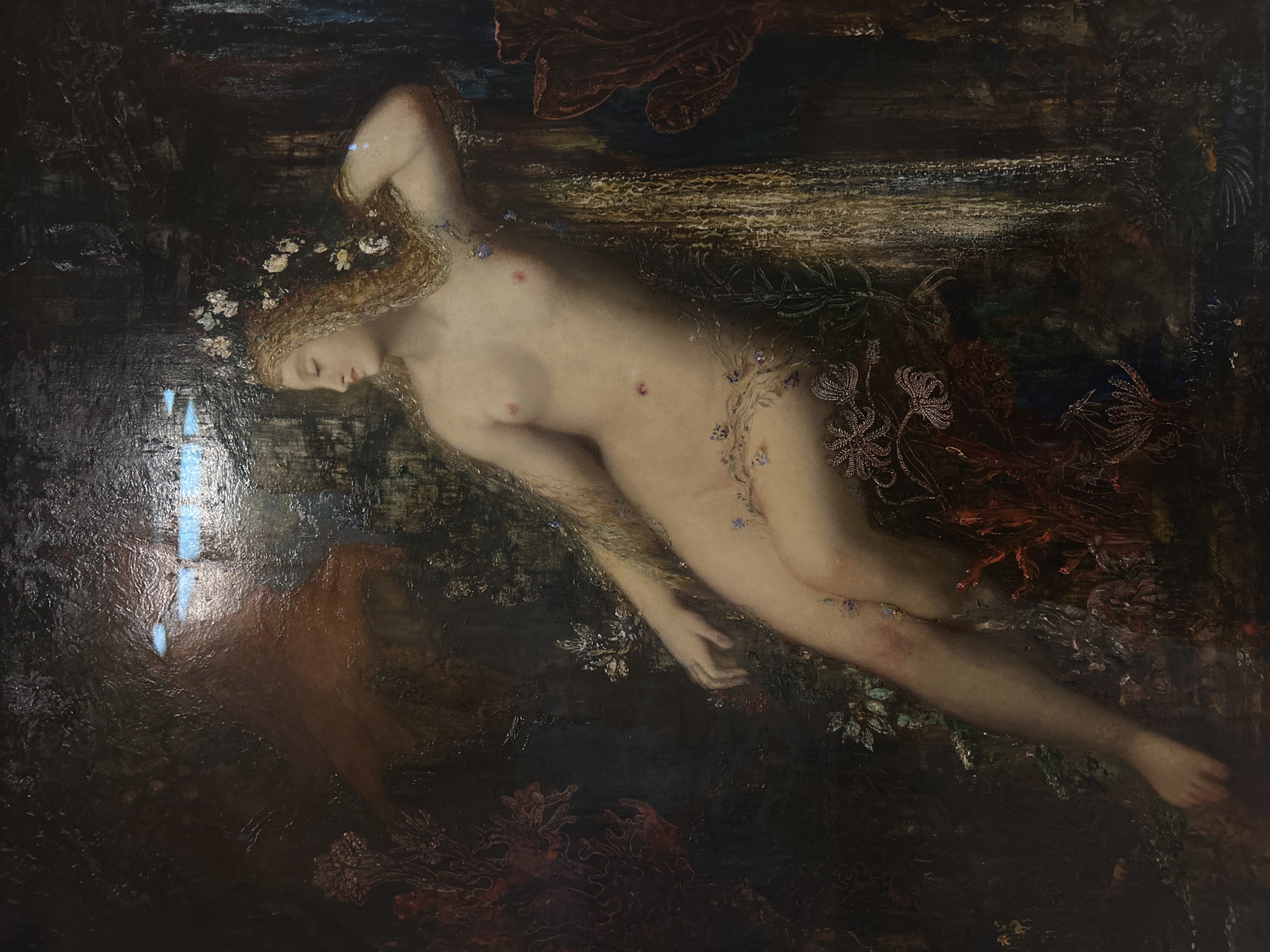 Oil painting depicting a reclining nude female figure with blonde hair and a floral wreath, surrounded by dark foliage. The scene takes place in an ambiguous, dreamlike environment with a glimpse of shimmering light. The painting is currently located in Paris, France.