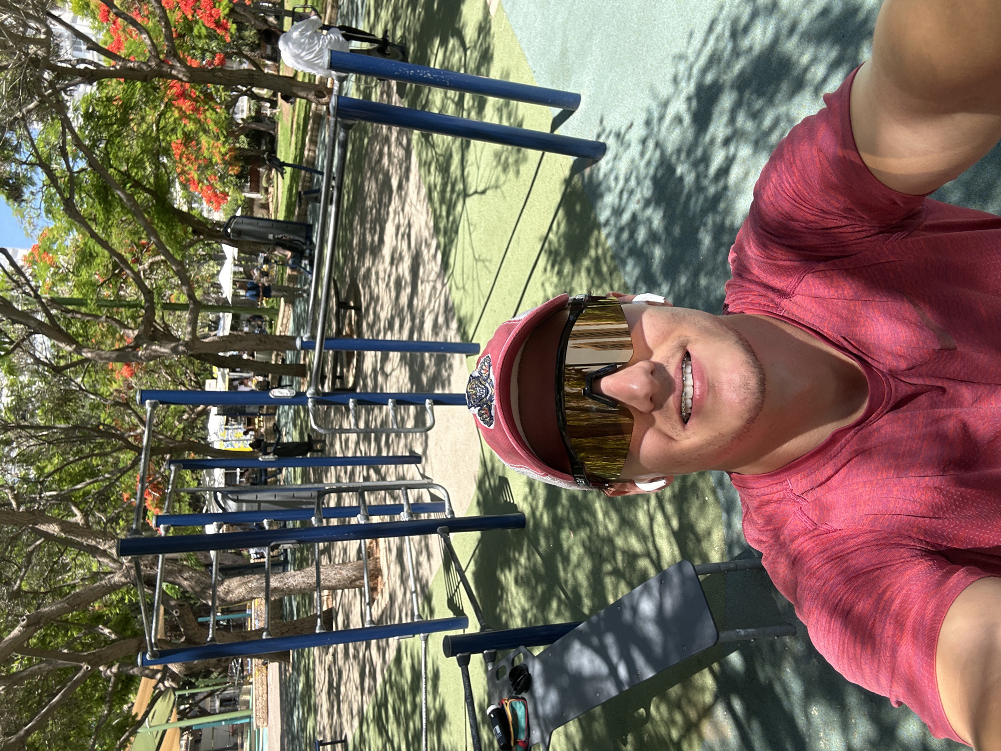 The image is a selfie taken by a young man wearing a red t-shirt, a red baseball cap, and large mirrored sunglasses. He has braces on his teeth and is wearing white wireless earbuds. He is standing in an outdoor fitness park with blue metal exercise equipment. In the background, there are trees with red flowers, other people using the equipment, and shaded areas with tables and umbrellas. The park has a green and beige rubberized surface. The scene appears to be in a public park in the city of Ramat Gan, Israel, given the presence of outdoor gym equipment common in Israeli city parks and the vibrant, sunny atmosphere.