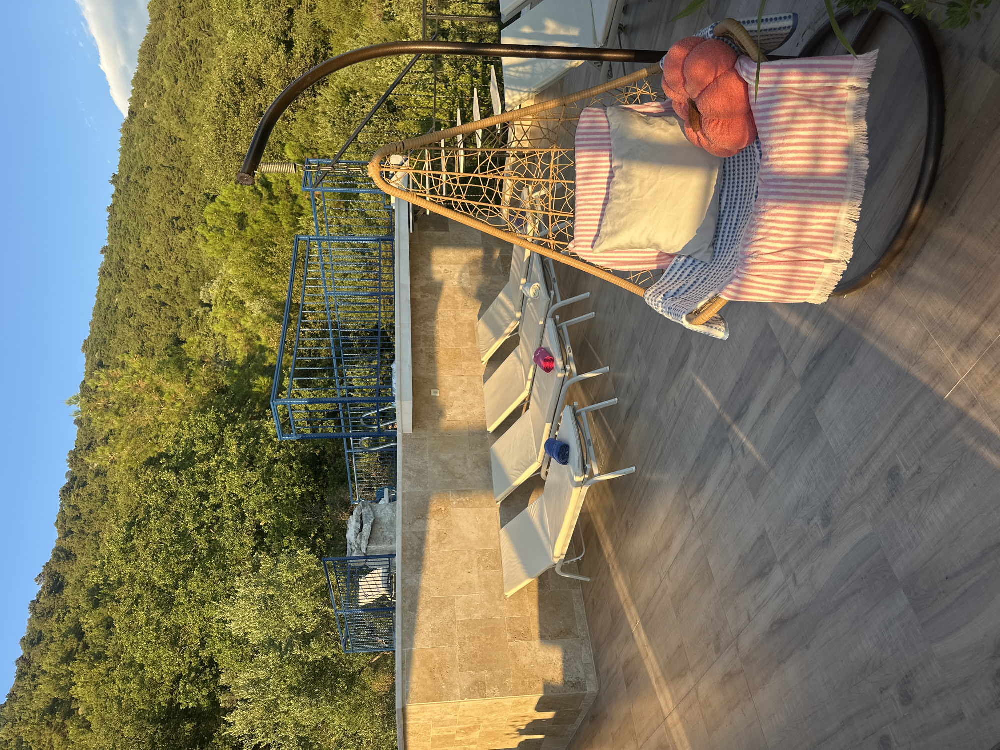 A tranquil outdoor seating area is depicted in Kavač, Montenegro, bathed in the warm glow of late afternoon sunlight. The scene overlooks a densely forested hillside, with lush green trees covering the slopes under a clear blue sky. In the foreground, a modern patio features a wooden-style tiled floor. Several beige lounge chairs with white frames are arranged neatly, each with a rolled blue towel placed on it. Adjacent to the lounge chairs, a unique hanging swing chair, made of woven rattan, is adorned with a striped pink and white blanket, a light-colored pillow, and a textured coral-colored cushion shaped like a pumpkin. Behind the seating area, a beige tiled wall serves as a backdrop, topped by a blue metal railing that encloses a raised terrace. Steps are visible leading up to this elevated area. The overall impression is one of relaxation and peaceful enjoyment of the natural surroundings.