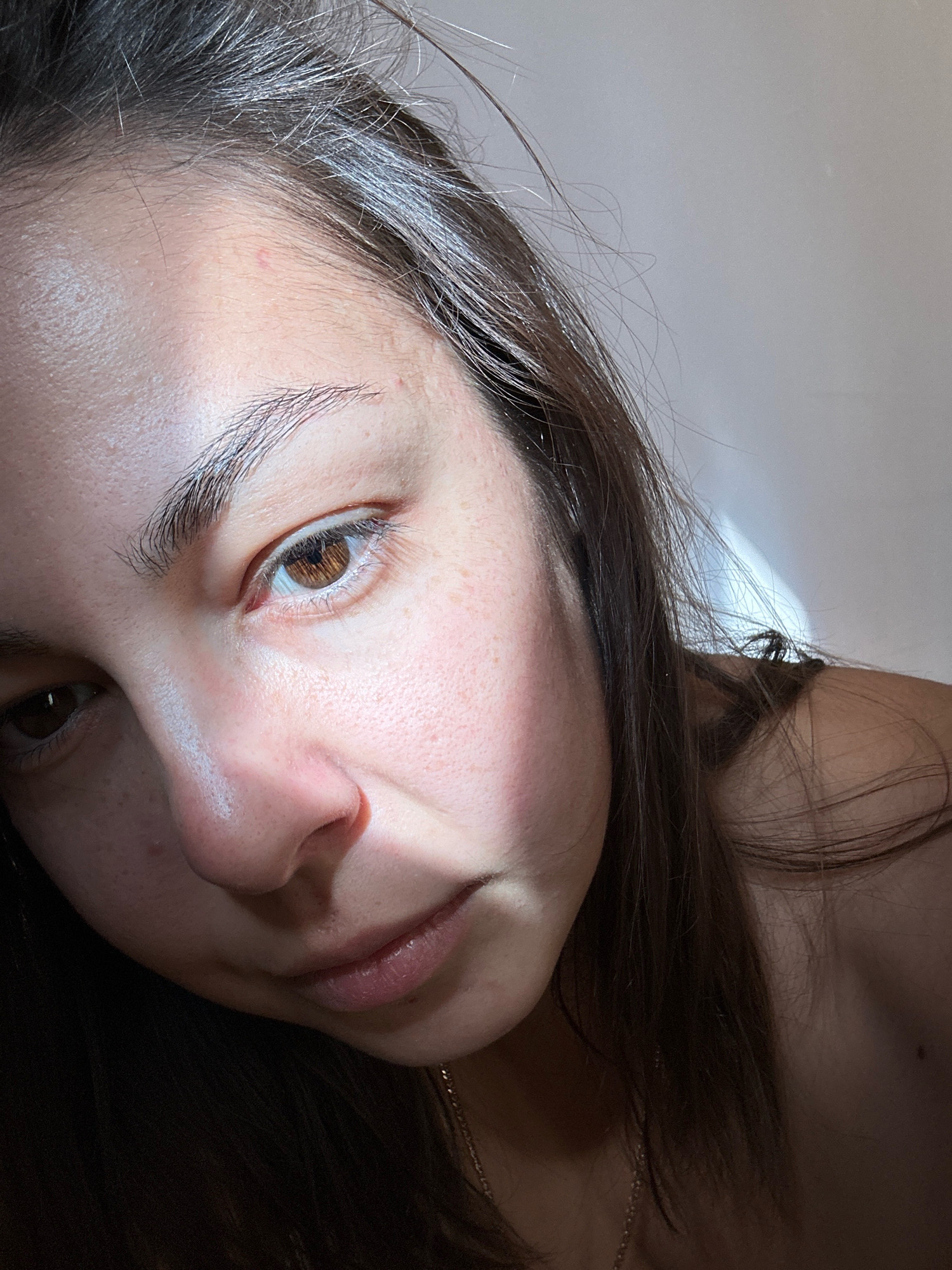 The image is a close-up, dimly lit portrait of a young woman's face, angled slightly away from the camera. Her brown eyes are visible, with one looking directly at the viewer and the other cast downwards. Her skin has a pale complexion with some freckles and subtle blemishes. The lighting casts a strong shadow across the right side of her face and highlights the left cheekbone and forehead. Her dark brown hair is loose and falls around her face and shoulders. A thin gold chain is visible around her neck. The background is largely out of focus and appears to be a neutral-colored wall. There are no discernible landmarks or elements that would indicate a specific location within the city of Gyumri, Armenia.