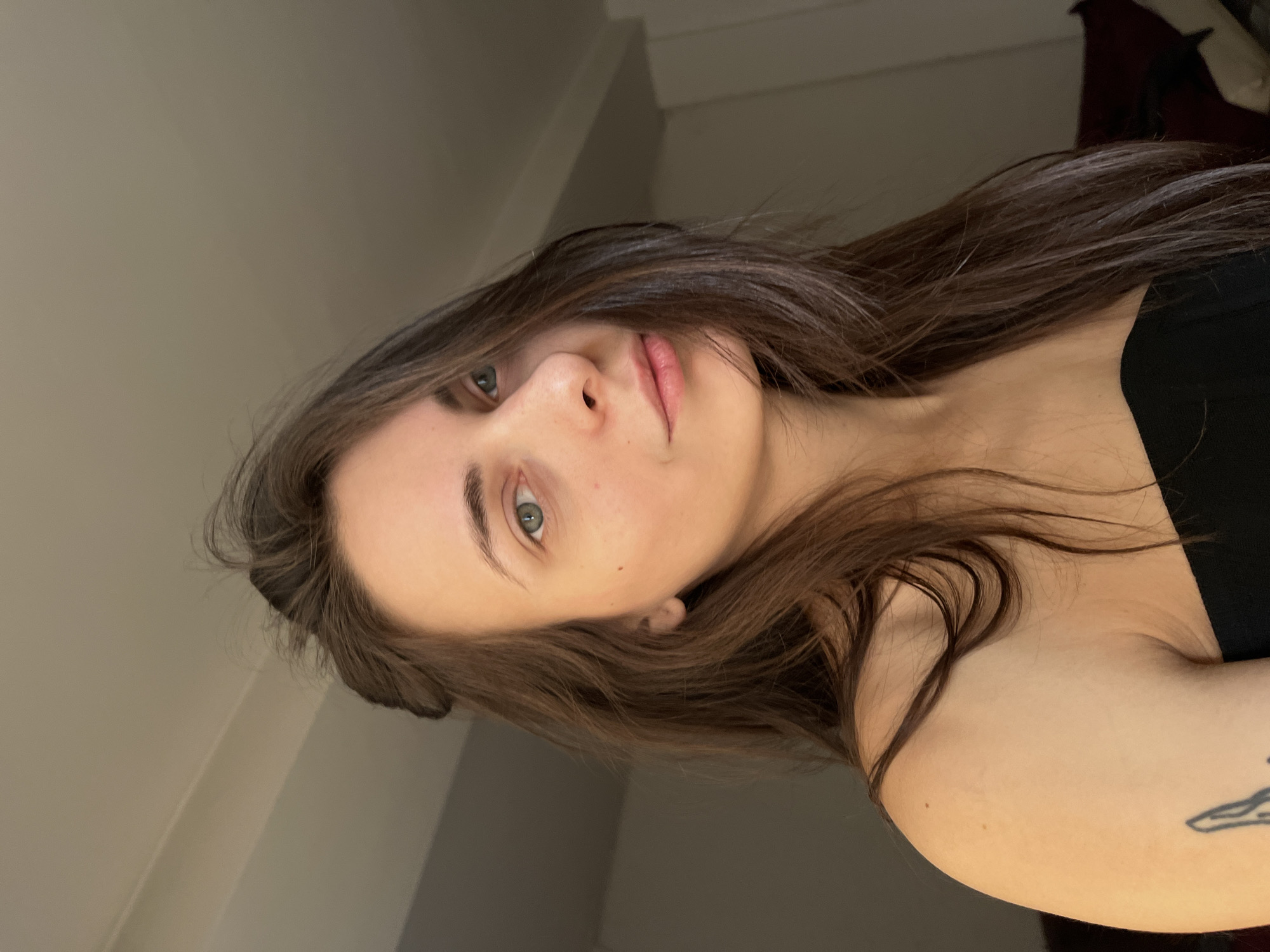 A portrait of a fair-skinned woman with long, wavy brown hair and green eyes. She is wearing a black top. A tattoo is visible on her left arm. The background appears to be an interior setting with light-colored walls and a glimpse of a dark curtain or closet. It is not possible to determine the location in Krasnoyarsk, Russia.