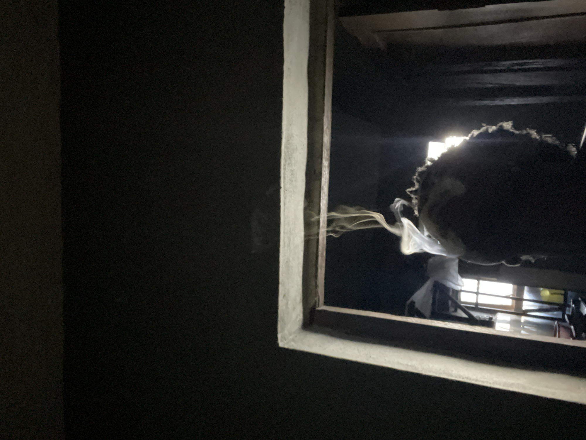 A low-angle, medium shot captures the silhouette of a person with curly hair, facing away from the camera and towards a doorway. Wisps of smoke rise from the top of their head, dissipating into the dark surroundings. The doorway frames a dimly lit interior where a window is visible, letting in a bright light that illuminates hanging fabric and other indistinct objects. The scene appears to be set within a room in Nkpor, Nigeria. The overall mood is somber and atmospheric.