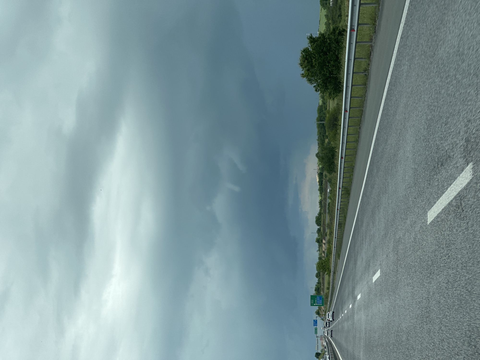 The image shows a highway in the city of Svoboda, Bulgaria. The road surface is gray asphalt, with white lane markings. A metal guardrail runs along the edge of the road. Beyond the guardrail, there is greenery and trees. A line of vehicles is visible in the distance. The sky is overcast with dark clouds.