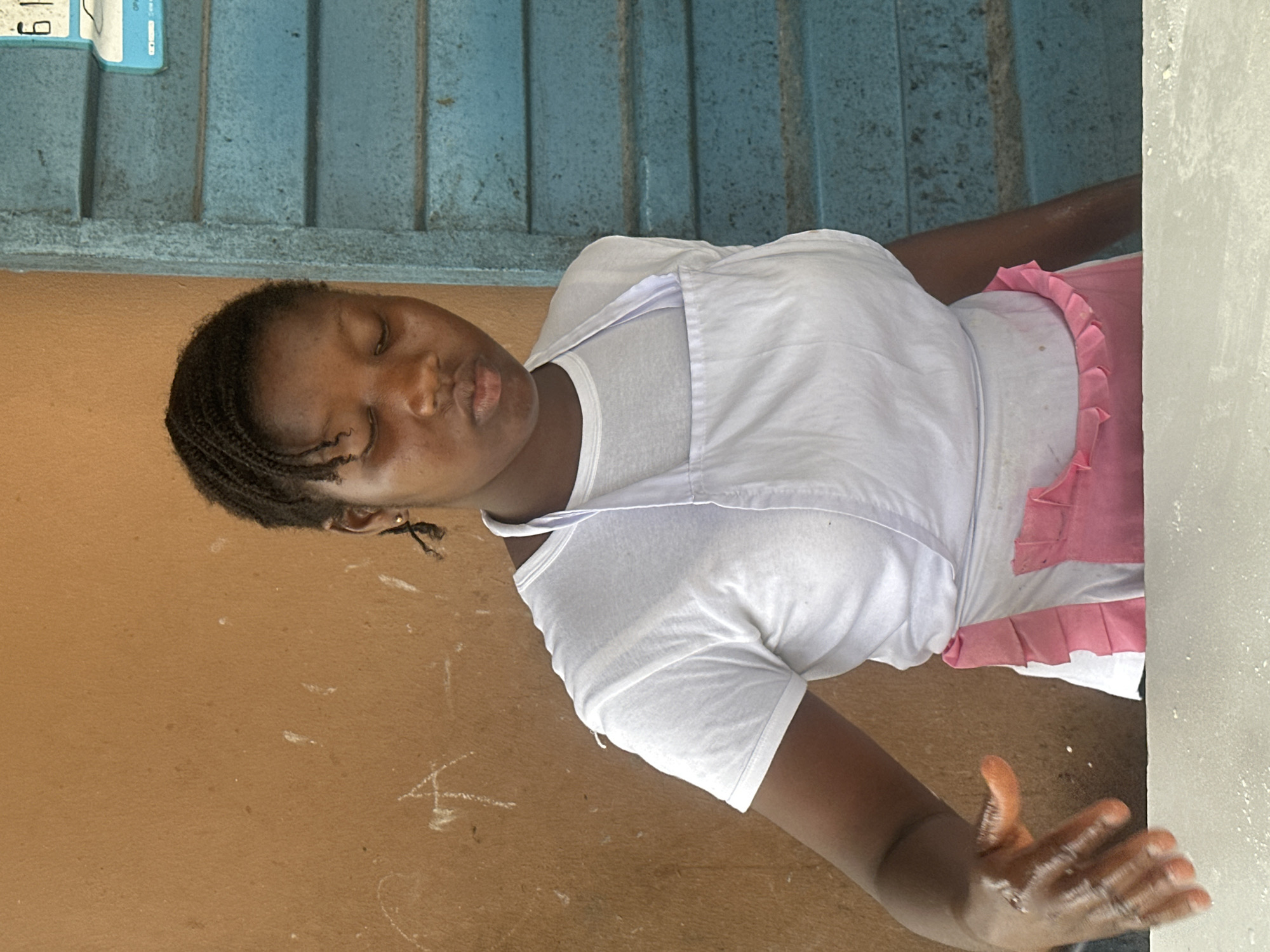A young woman with braided hair is standing and looking down, her eyes closed. She is wearing a white t-shirt and a white apron with pink ruffles. Her left hand is in motion, appearing to be touching or working with something on a white surface in the foreground. The background consists of a textured, reddish-brown wall on the left and a blue, slatted structure on the right, which has a visible price tag or label with the number "61" at the top. The scene is likely outdoors or in an open-air market in Badagry, Nigeria.
