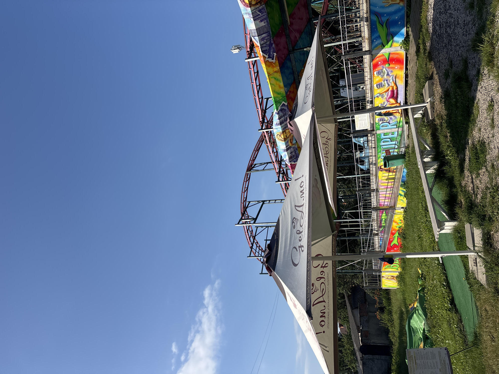 The image shows an outdoor amusement park scene on a clear, sunny day. In the foreground, two large, white umbrellas with "Gelato Amore" written on them provide shade. Beneath the umbrellas, there is a white bench and some grassy areas with a small putting green. Behind the umbrellas, a brightly colored amusement park ride with a roller coaster track is visible. The ride features a vibrant mural with images of people and cityscapes. A small building and trees are visible in the background on the left side of the image. The sky is a bright blue with a few scattered white clouds.