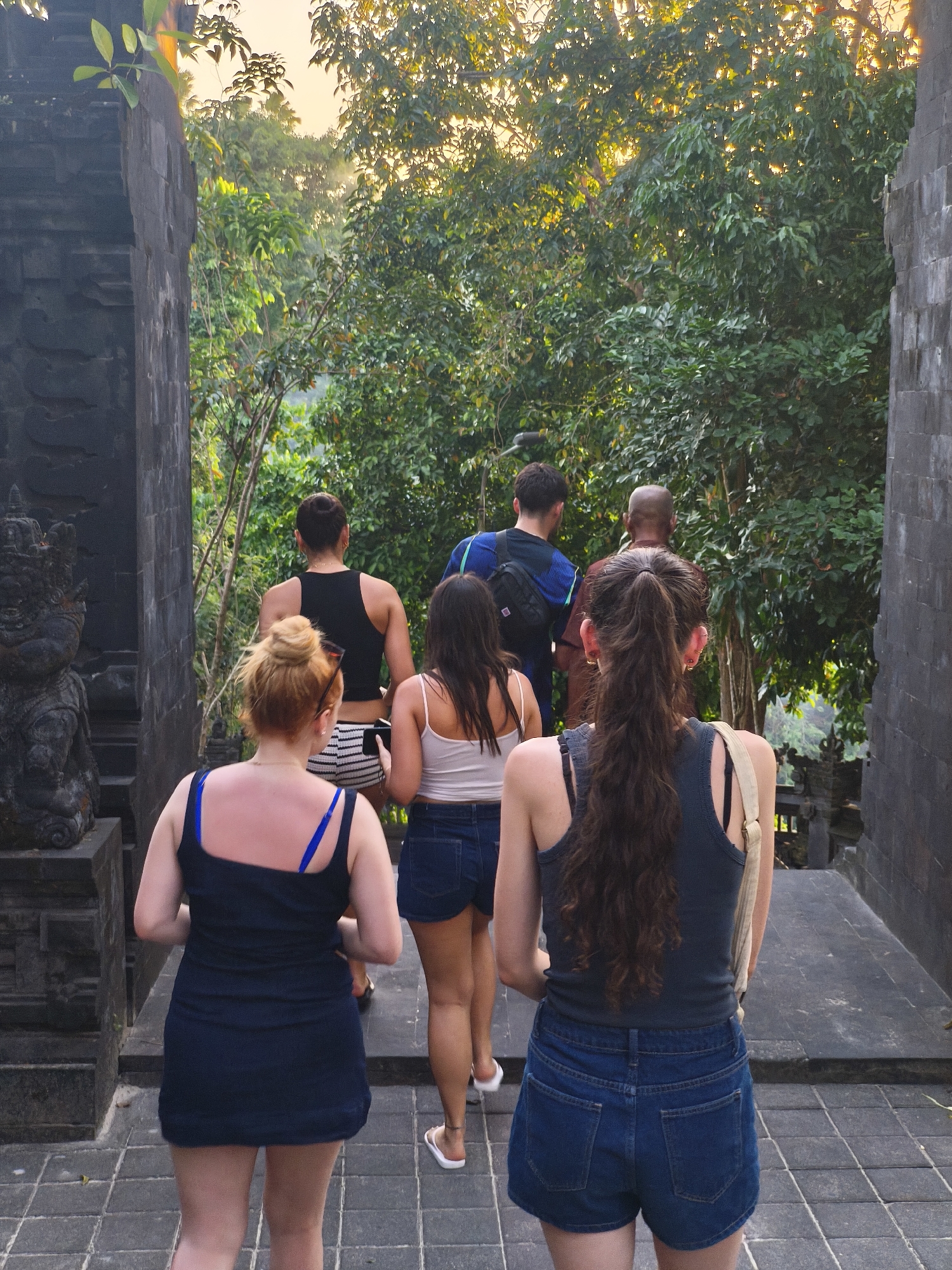 In Kuta Selatan, Indonesia, a group of six individuals walks up a stone staircase towards a lush green tree-lined pathway. The scene is framed by dark stone structures on either side, one featuring a carved statue on the left. The group consists of four women and two men, all walking away from the camera. The women are in the foreground and middle ground, wearing casual summer attire including shorts and tank tops. The men are further up the stairs, one in a blue shirt and backpack, the other in a reddish-brown shirt. The environment suggests an outdoor temple or historical site, with sunlight filtering through the dense foliage overhead, creating a warm, natural ambiance.