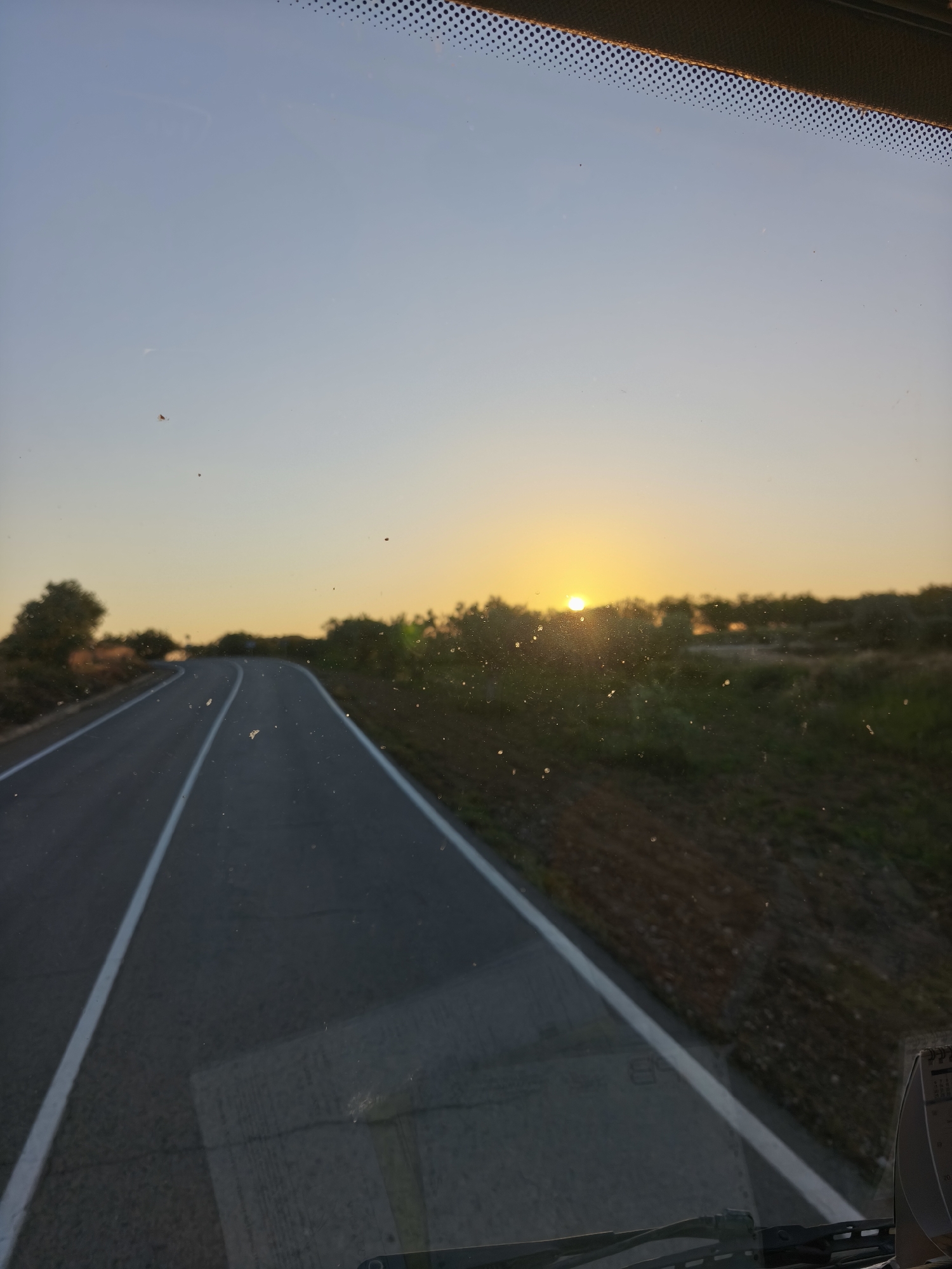 The image, likely taken from inside a vehicle moving along a paved road, presents a view of a rural landscape at what appears to be sunrise or sunset. The road is marked with solid white lines and curves slightly to the left in the distance. Along the roadside, there are trees and vegetation. The sun is visible, low on the horizon, slightly obscured by the foliage. The sky is a gradient of light blue to yellow-orange. The image is slightly obscured by what seems to be dirt or spots on the windshield. The location is possibly in the vicinity of Turís, Spain, due to its rural character and the presence of a highway.