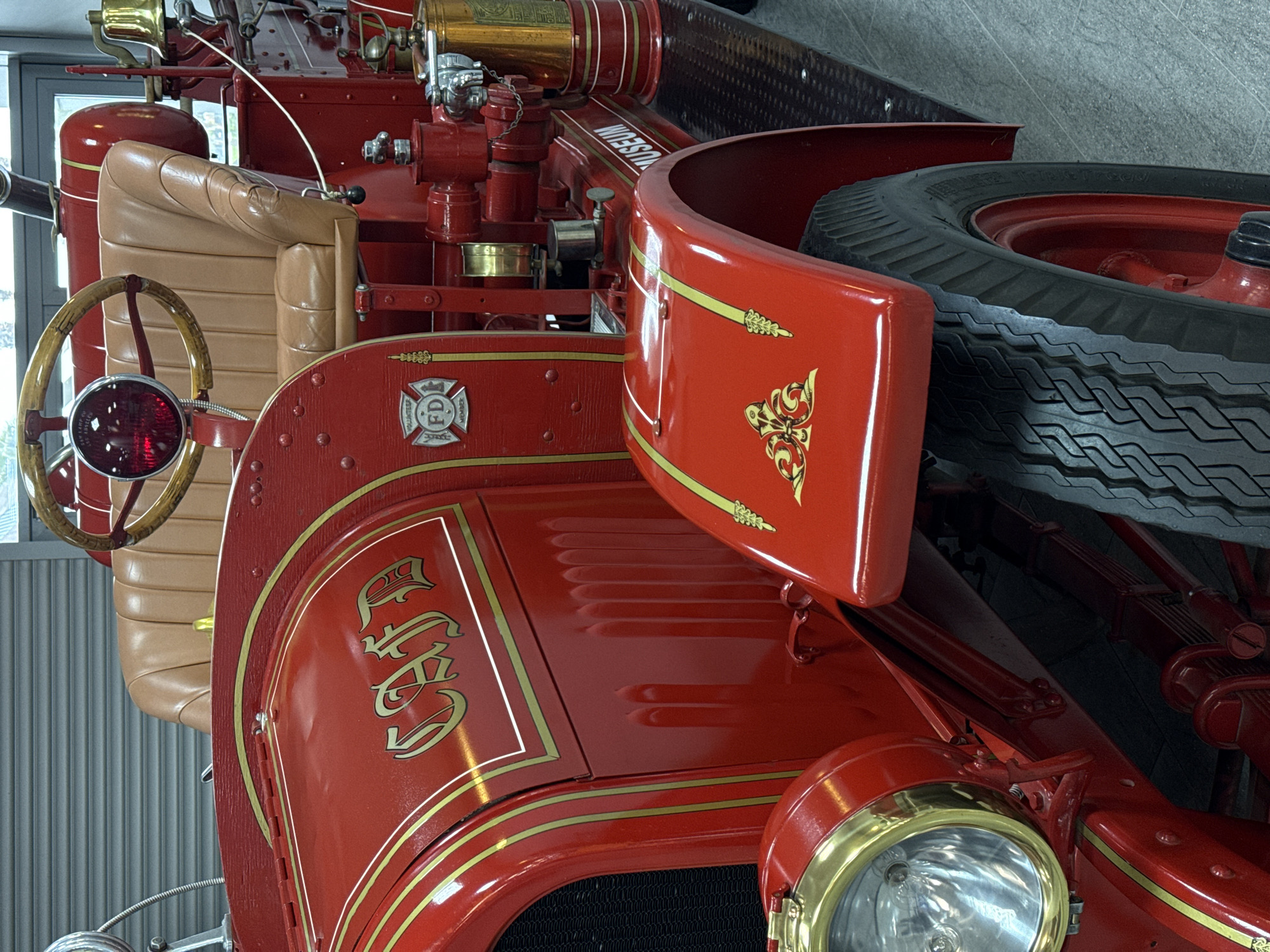 The image shows a section of an antique fire truck. Prominently displayed are the red body panels with gold trim, the running board with a spare tire, a tan leather seat, and a wooden steering wheel. The fire truck has a headlight with a brass trim ring. There is a badge that has the letters F and D with the words VOLUNTEER and MEMBER around them. In addition, the letters "CSFD" are emblazoned on the side. The location is indeterminable from the photo. It is impossible to know from this image if the photo was taken in Sochi, Russia.