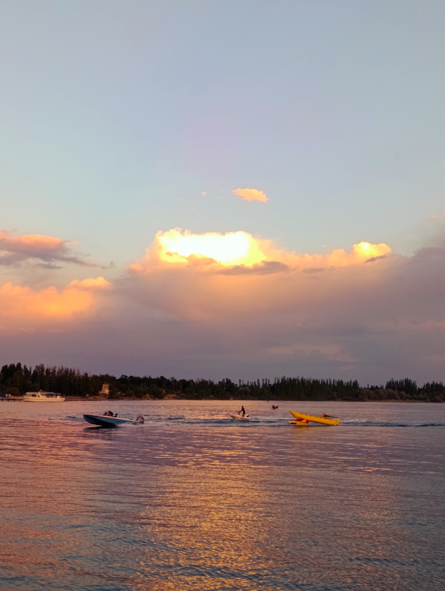 The image depicts a serene sunset over a body of water, likely Issyk-Kul Lake, in Bayet, Kyrgyzstan. The sky is painted with soft hues of pink, orange, and blue, with scattered clouds catching the last rays of sunlight. A line of trees forms the horizon, with a white boat docked on the left. In the water, a speed boat is seen in motion, leaving a wake. Further out, a yellow kayak or a similar watercraft is also visible. The water reflects the colors of the sky, creating a shimmering effect. The overall atmosphere is tranquil and picturesque, capturing a moment of natural beauty at dusk.