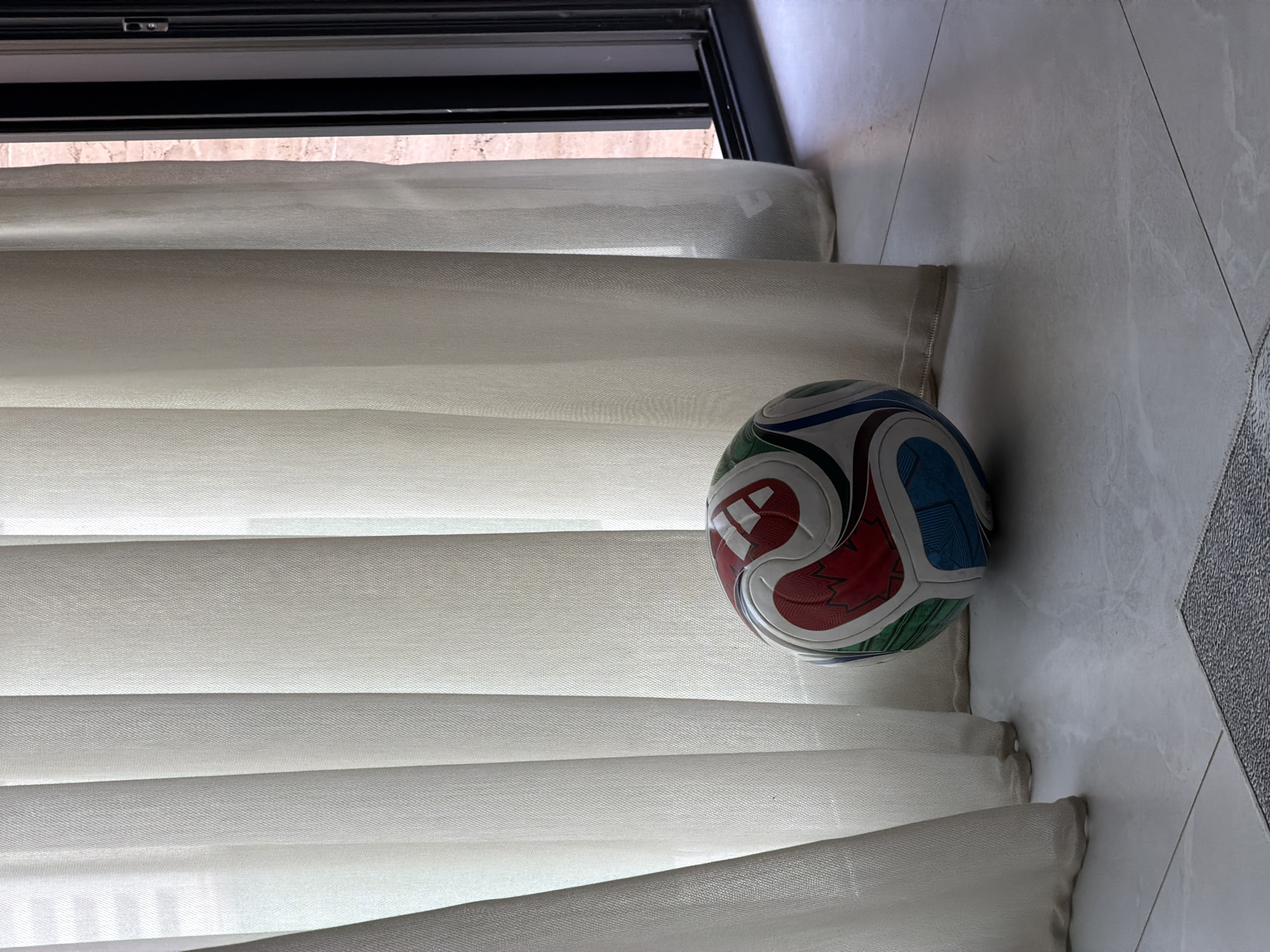 A soccer ball is resting on a pale gray tiled floor in front of sheer white vertical blinds. The ball is multi-colored with red, white, blue, and green panels. To the right of the blinds, a dark window frame is visible, with a glimpse of a reddish-brown brick wall outside. The scene appears to be indoors, possibly in a residential setting in Finestrat, Spain, characterized by modern architecture and natural light filtering through the blinds. No people are present in the image.