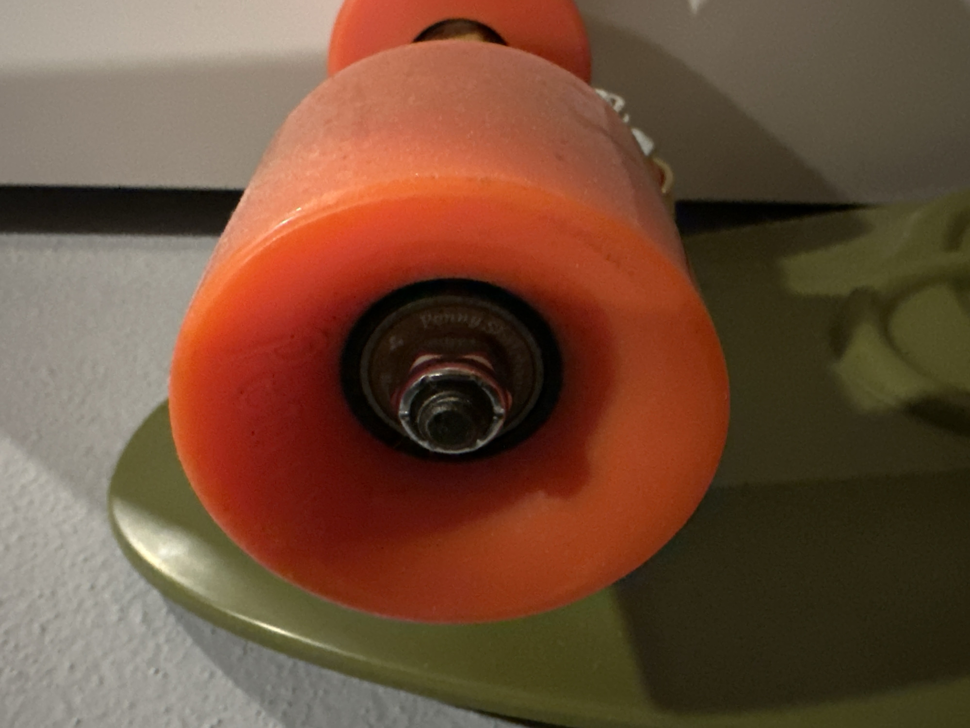 A close-up photograph shows a portion of a penny board skateboard. The skateboard is olive green and has orange wheels. The wheels are dusty. A metal bearing is visible within the center of the wheel. The surrounding environment is a light gray wall. The photo was taken indoors. The location is unknown and cannot be determined from the image.