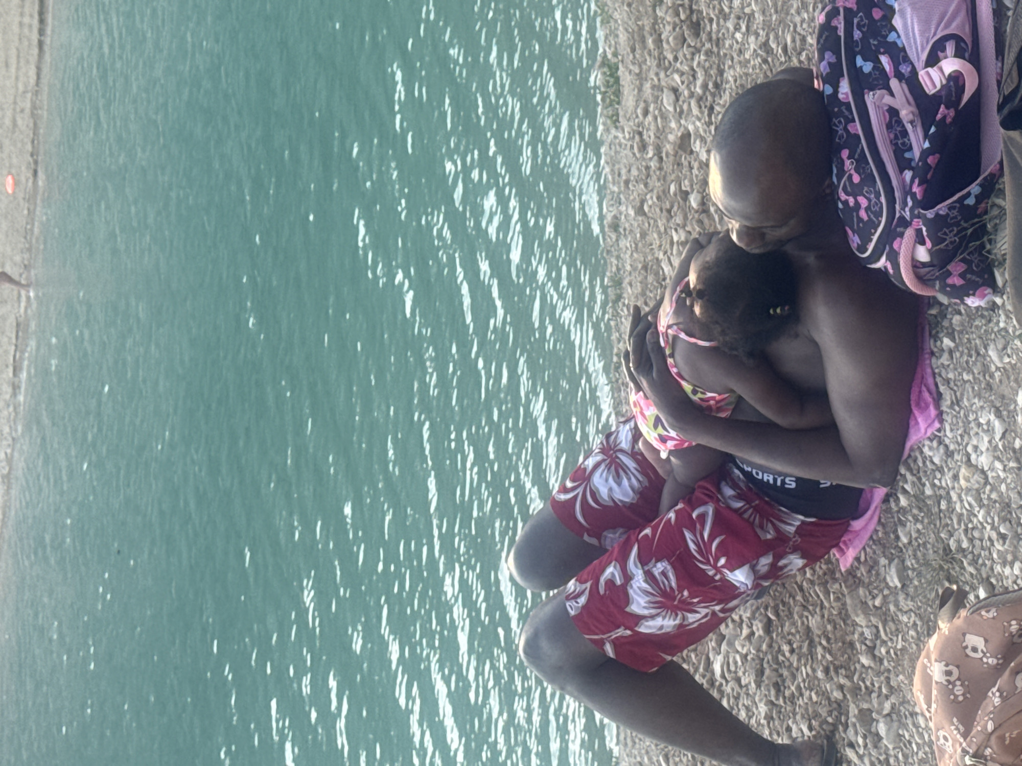The image shows a man holding a young child close to him while sitting on a rocky shore next to the water. The man is wearing red swim trunks with a white floral pattern and black underwear with "SPORTS" written on them. The child is wearing a pink and yellow patterned swimsuit. A backpack with a butterfly pattern is visible behind the man. The water is a vibrant teal color, reflecting the sunlight. The scene appears to be during the daytime, with bright light casting shadows. The rocky shore suggests a natural outdoor setting. The man's embrace of the child conveys a sense of warmth and affection, hinting at a peaceful or loving moment between them. Another person is partially visible in the lower right of the image, wearing a patterned garment.