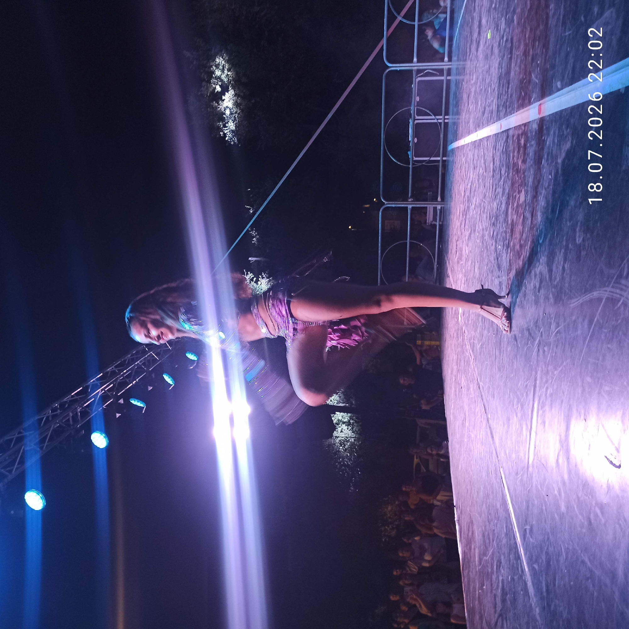 An adult female performer, wearing a sparkling purple costume and high-heeled sandals, is captured mid-dance on an outdoor stage at night. She is positioned on the right side of the frame, with her left leg raised and her body angled to the left. Her face is turned to the left, and she appears to be smiling. Bright, blurred blue and purple stage lights illuminate the scene, creating streaks of light across the image. The stage floor is a dark, reflective surface. In the background, a crowd of spectators is visible, along with trees and a metal railing. The scene is located in Bitonto, Italy, as indicated by the timestamp "18.07.2026 22:02" which suggests a date and time for the event.