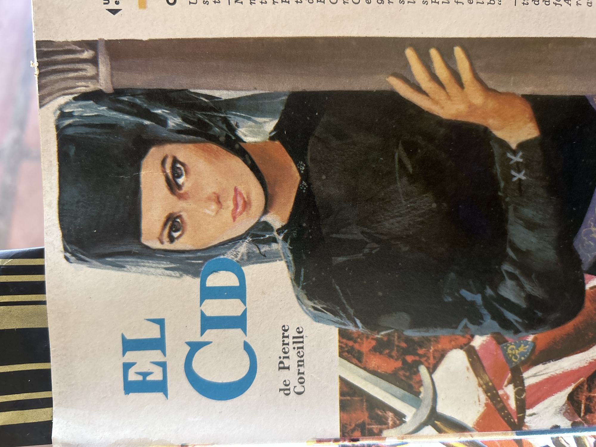 The image is a close-up of a magazine cover featuring a painting of a woman in historical attire. The woman wears a black headdress that covers her hair and a black garment. She has dark hair, expressive eyes, and is looking directly at the viewer. The text on the cover reads "EL CID" in large blue letters, followed by "de Pierre Corneille" in smaller black letters. To the left of the woman, a portion of a sword and a shield with a crest are visible, suggesting a theme of historical drama or epic. A column is present on the right side of the image. The background appears to be a textured surface. The scene is static and artistic, not a real-world event. This image does not provide any information about the city of Santa Ana, Venezuela.
