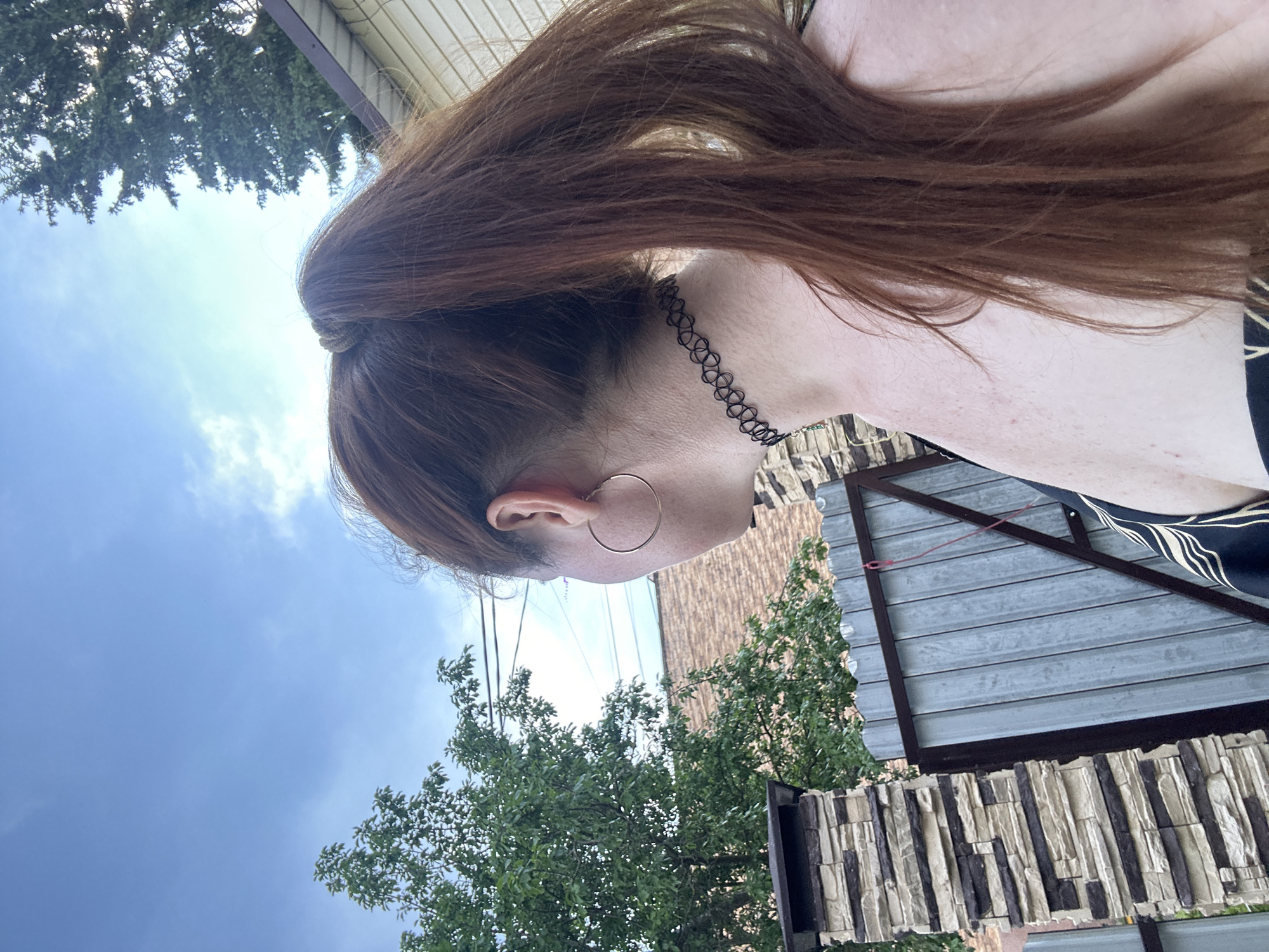 A woman with auburn hair pulled into a ponytail is shown from the back and side, wearing a black choker necklace and a hoop earring. She is outdoors, with a cloudy blue sky and green trees in the background. To her left is a stone pillar and a corrugated metal fence. The scene appears to be a backyard or residential area, possibly in Nizhny Novgorod, Russia, given the architectural style of the house visible in the background.