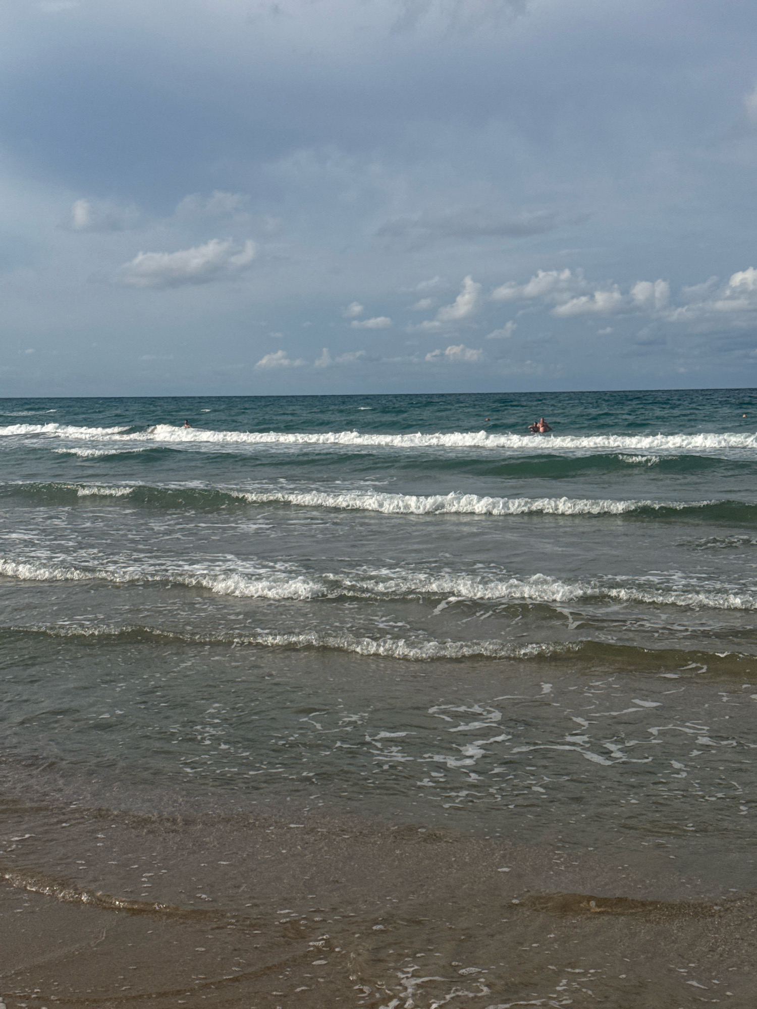 A wide shot captures the gentle waves of the Black Sea lapping at a sandy beach in Anapa, Russia. The water is a deep turquoise, with white foam cresting on the waves. In the distance, two individuals are visible in the water, appearing to be swimming or wading. The sky above is partly cloudy, with a mix of light and dark gray clouds against a pale blue backdrop. The sand on the beach is a muted brown, with a slight sheen from the water. The scene is serene and typical of a summer day at the seaside.