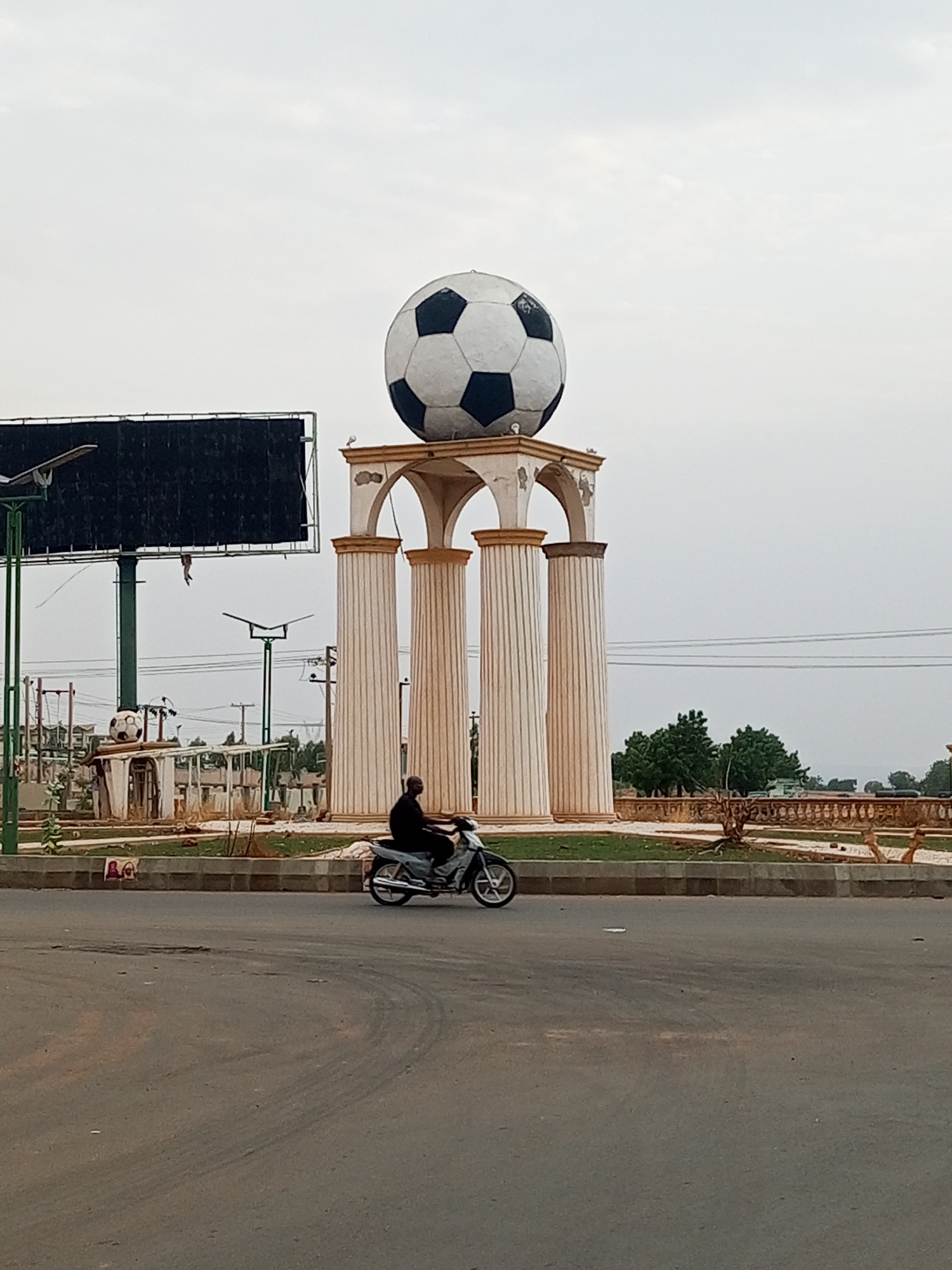 The image depicts a roundabout in Gombe, Nigeria. At the center of the roundabout is a monument consisting of four faux-Corinthian columns supporting a large soccer ball. A motorcyclist rides on the road in front of the monument. There is a large billboard on the left of the image. The sky is overcast.