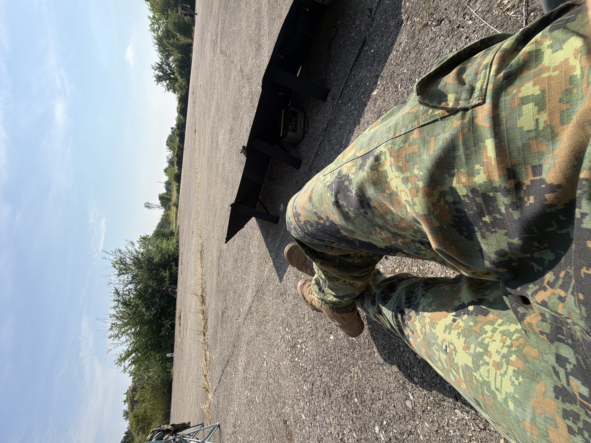 The image depicts an outdoor scene in Bogdanitsa, Bulgaria, observed from a first-person perspective. The primary subject's legs, clad in German Flecktarn camouflage pants and tan tactical boots, are extended across the foreground, resting on a cracked, grey asphalt or concrete surface. To the subject's right, a black foldable solar panel array is deployed on the ground, powering a Goal Zero Yeti-style portable power station visible beneath it.
The immediate ground surface is sparsely covered with dry grass and weeds. In the midground, the paved area extends towards a line of dense green bushes and small trees. Beyond these, additional sparse trees and distant foliage are visible under a clear, bright blue sky with scattered white clouds. Strong shadows indicate a sunny environment.
In the left background, a light blue folding chair with camouflage-patterned gear draped over it is present. Further to the left, behind vegetation, a figure is partially discernible on higher ground. Another dark figure is visible near the base of the bushes in the center-right background, and a plastic water bottle rests on the ground nearby. On the far right edge of the background, a lone individual is observed standing or walking. The overall scene suggests an outdoor activity or rest period in a semi-rural or abandoned paved area.