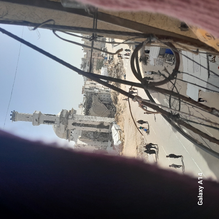 An elevated view captures a scene of extensive structural damage in Khan Yunis, Palestinian Territory, under a clear, bright sky. The prominent structure in the mid-ground left is a heavily damaged white mosque, identified as the Great Mosque of Khan Yunis. Its minaret stands partially intact but shows significant pockmarks and damage, with visible rebar and crumbling sections. The main body of the mosque is in a state of partial collapse, with large sections of walls missing, exposing interior rubble and twisted metal.
The area immediately surrounding the mosque and extending into the background is a rubble-strewn landscape, indicating widespread destruction. Several other buildings in the background also display varying degrees of damage or collapse. Makeshift shelters, constructed from white tarpaulins, are visible among the debris near the base of the mosque.
A paved asphalt road traverses the mid-ground, serving as the primary pathway for numerous pedestrians. Individuals are observed walking in both directions along the road. On the left side of the road, a group of people, some in dark, traditional attire, walk together; one individual appears to be pushing a white cart or similar wheeled object. Further down the road, another person pushes a blue cart. Other scattered individuals and small groups walk across the scene.
The image is framed by foreground obstructions, including a large, blurry dark mass on the left, and a network of dark, rusty metal bars and wires, possibly part of a balcony railing or window frame, on the right and across the upper mid-ground. A blurry pinkish object is visible on the far right.