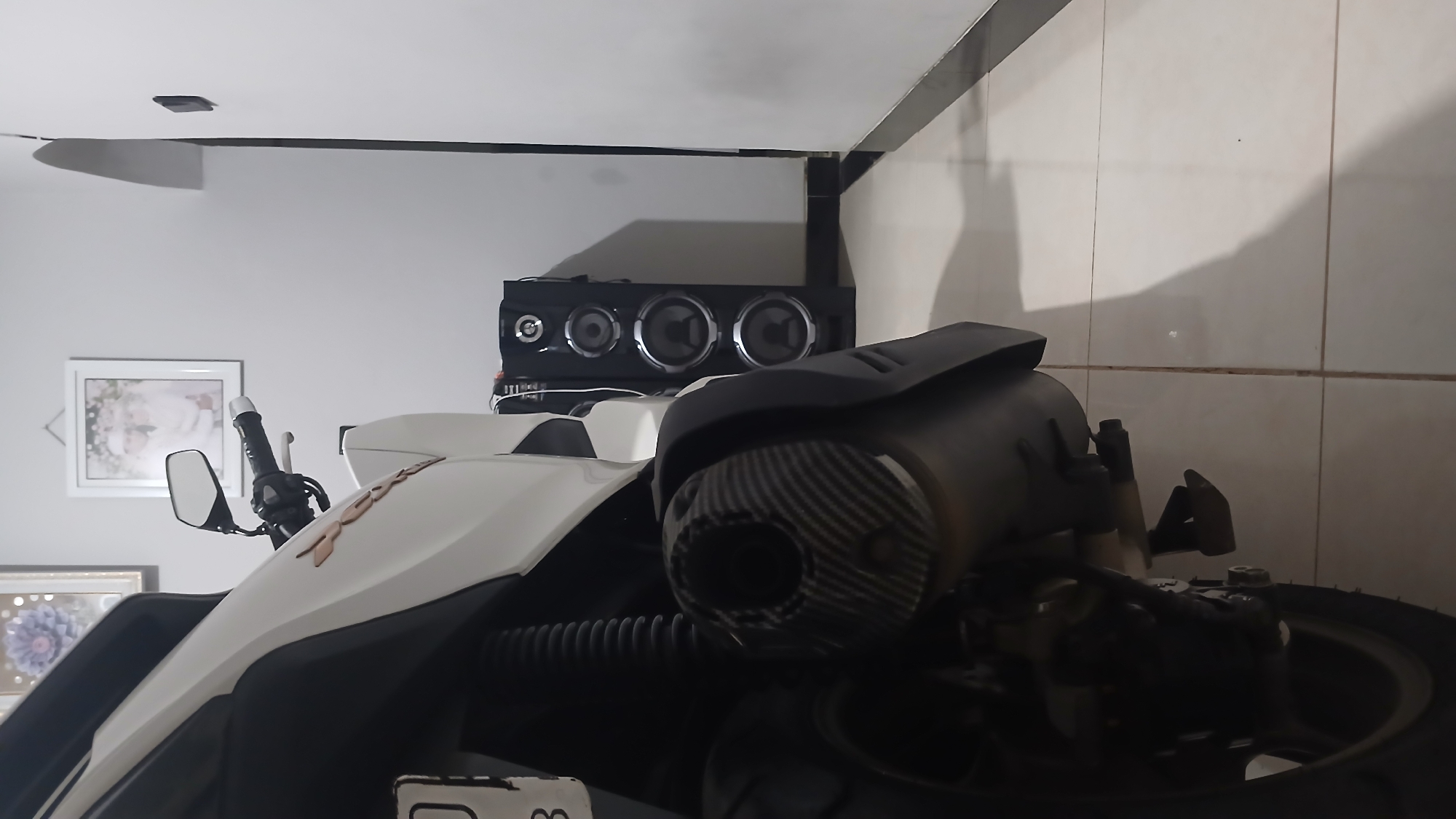 The image is a low-angle shot taken indoors, likely in Sampang, Indonesia, focusing on the rear section of a white and black motorcycle. The motorcycle's exhaust pipe, which has a carbon fiber pattern, is prominent in the foreground. Behind the motorcycle, a tall, black speaker system with multiple drivers is visible against a light grey wall. To the left of the motorcycle, part of a license plate with "83" is visible. Higher up on the wall, two framed pictures are hung: one on the left with a floral design and another on the right containing a portrait of a couple. The floor is tiled with light-colored square tiles. The lighting appears to be artificial and somewhat dim, casting shadows on the floor. There are no people or animals present in the image.