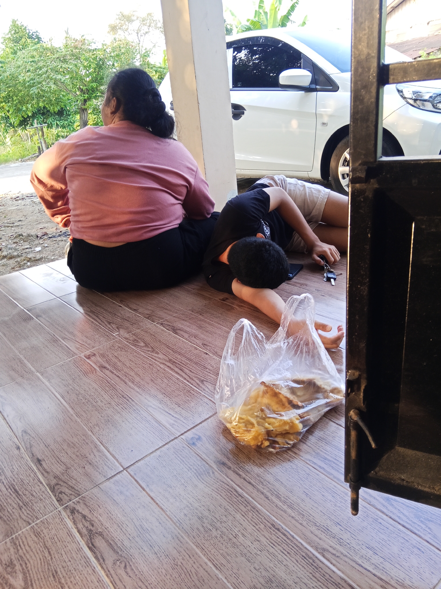 The image depicts an outdoor scene on a tiled patio, likely in Lubuklinggau, Indonesia, as evidenced by the presence of a white car and tropical foliage. In the foreground, a person is lying on their back, asleep, with a plastic bag containing what appears to be food beside them. A set of keys rests on the ground near the person's hand. To the left, another person is seated with their back to the camera, also appearing relaxed or asleep. The background shows a street and lush green trees, suggesting a residential or semi-rural setting. A portion of a black metal structure, possibly a gate or fence, is visible on the right side of the frame. The lighting indicates it is daytime.