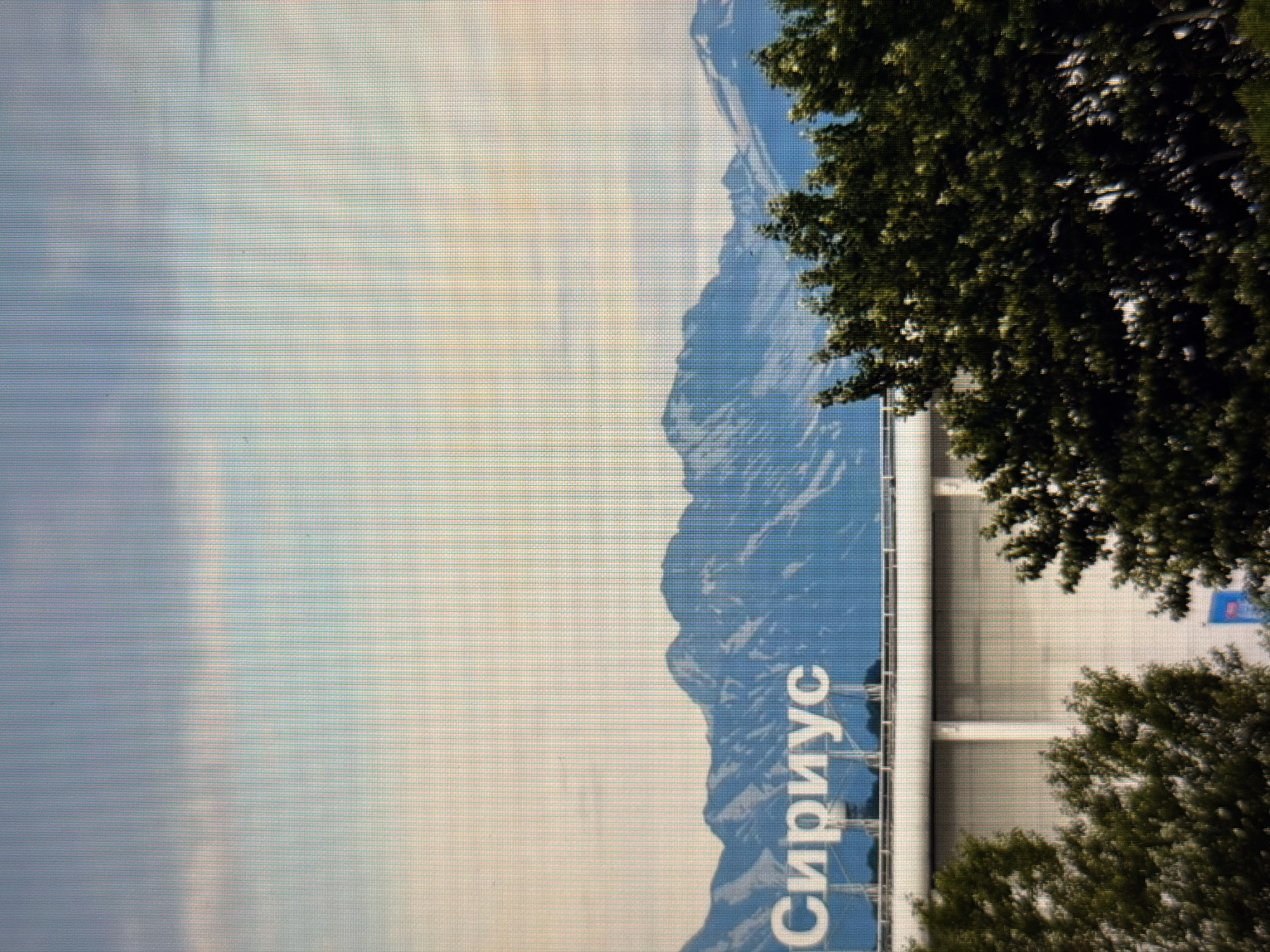 The image captures a wide shot of the city of Sirius, Russia. In the foreground, lush green trees partially obscure a large, white, modern-looking building. Above the building, a prominent sign displays the word "Сириус" in large, white, sans-serif lettering, set against a backdrop of majestic, blue-tinged mountains. The sky above is a blend of pale blue and white, with soft clouds scattered across it. The overall impression is one of a bright, clear day.