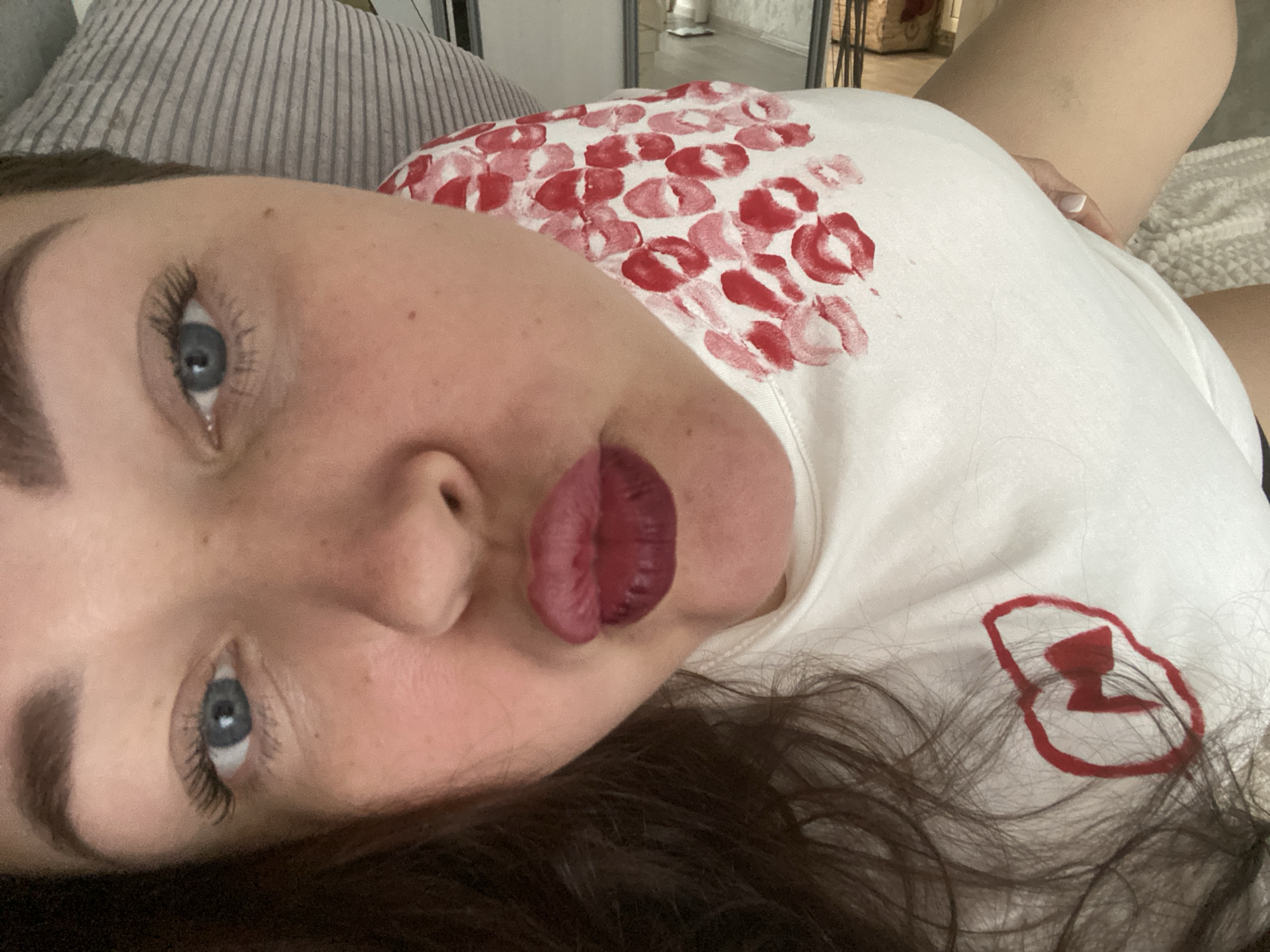 A woman lies on her side, resting her head on a striped pillow. She has fair skin, blue eyes, and dark eyebrows and hair. Her lips are painted a deep red. She wears a white T-shirt adorned with red lipstick prints on the upper part and a red logo on the lower right side. The background includes a light-colored wall with a glimpse of a mirror reflecting indoor objects. The scene provides no information related to the city of Sochi, Russia.