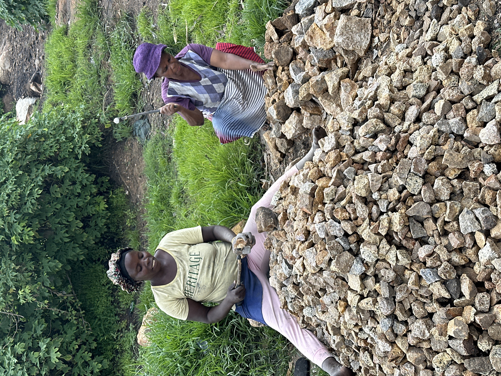 Two women are seen working with stones in an outdoor environment. The woman in the foreground, on the left, is wearing a yellow t-shirt with the word "HERITAGE" printed on it, a blue wrap around her waist, and pink trousers. She is sitting with her legs spread out and is actively engaged in breaking a stone with a hammer and chisel. A large pile of broken stones is in front of her.
The second woman, on the right, is wearing a purple headscarf and a checkered bib over a purple shirt, along with a striped skirt. She is also crouched and appears to be breaking stones, holding a metal tool in her hand. She is positioned behind another pile of stones.
The background consists of lush green vegetation, with trees and bushes, and some rocky terrain. The ground appears to be dirt and scattered debris. The lighting suggests it is daytime. The location is identified as Gura Top, Nigeria.