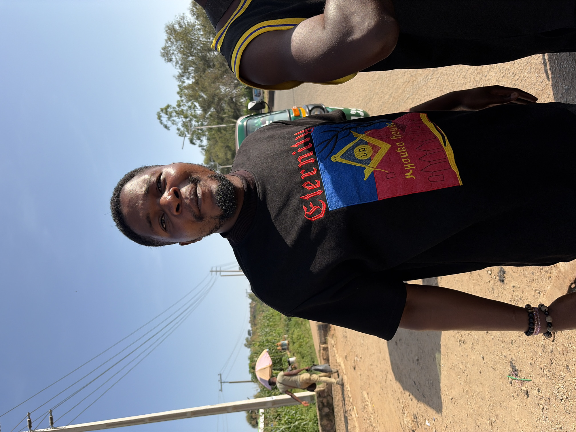 The image shows a medium shot of a Black man standing outdoors, possibly in Gura Top, Nigeria, on a sunny day. He is wearing a black t-shirt with a colorful emblem on the chest that includes the word "Eternity" and a symbol with "LD" inside a square and compass. He has a beard and is looking towards the camera. To his right, another Black man is partially visible, wearing a black tank top with yellow trim. In the background, there is a dirt road, vegetation, power lines, a utility pole, and a small green vehicle. Further in the distance, a person with an umbrella is walking along the road.