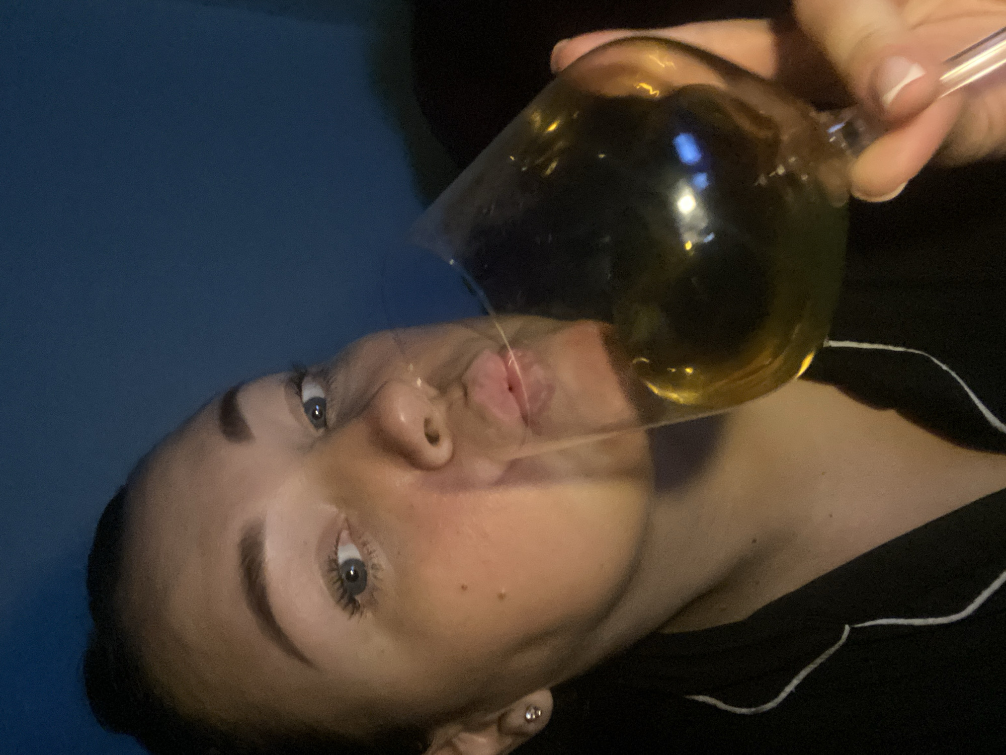 A woman is drinking from a large stemmed glass containing a yellow liquid. The glass is tilted towards her face, and a thin stream of liquid is flowing from the glass towards her mouth. She is wearing dark pajamas with white piping. The background is dark and blurred, possibly a wall. The location in Sochi, Russia, cannot be determined from the image.