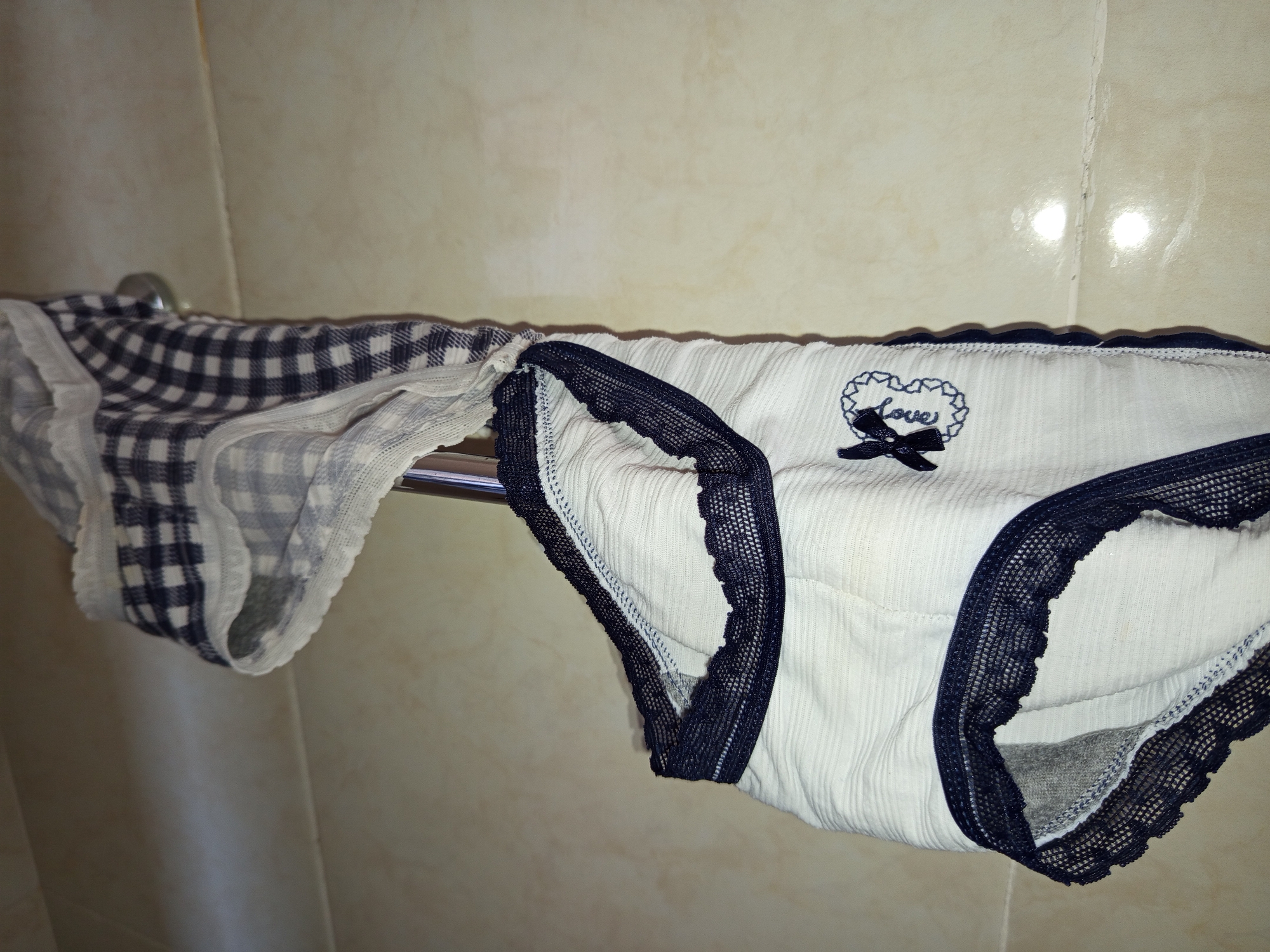 Two pairs of women's underwear hang on a metal bar against a tiled wall. The pair on the left is a blue and white checkered pattern. The pair on the right is white with dark blue lace trim and a small embroidered "love" design with a bow. The location is unspecified, but the city is North Kuta, Indonesia.