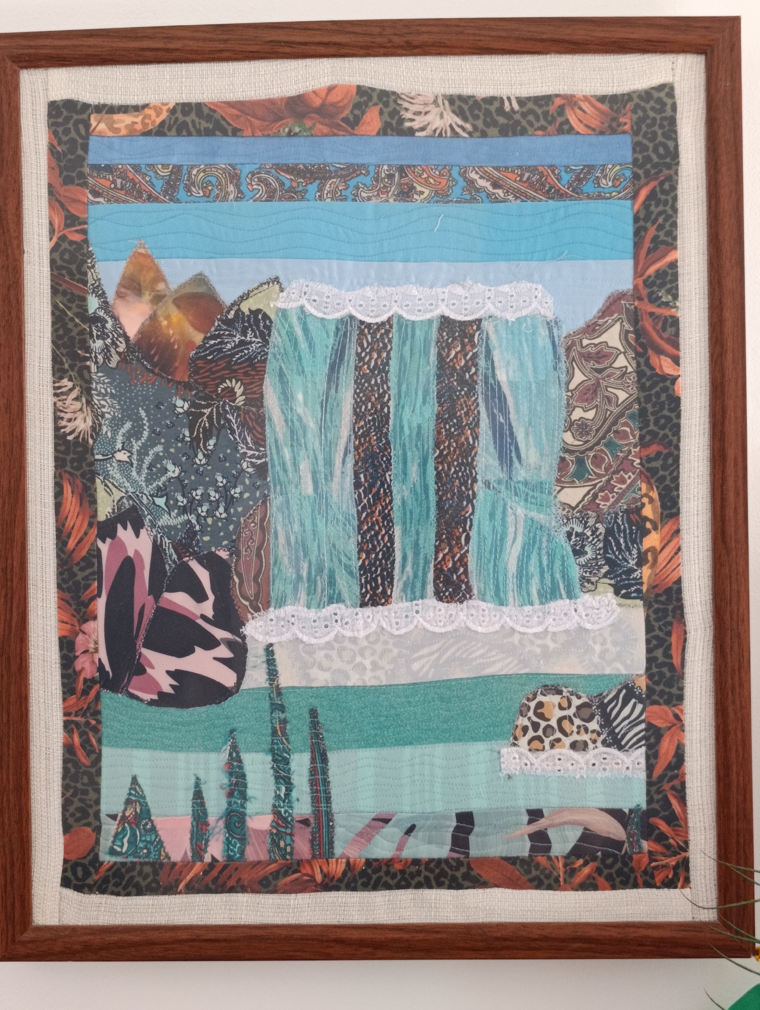 The image shows a framed textile collage. The collage is composed of various fabric scraps arranged in horizontal and vertical strips, depicting a stylized landscape. At the top, there are bands of fabric with patterns resembling paisley and leopard print, interspersed with solid blue and light blue stripes. Below these, a layered section features what appears to be mountains or hills made from patterned fabrics. The central portion of the collage depicts a waterfall or draped fabric in shades of turquoise and teal, with vertical brown and black stripes. This is bordered by white lace trim. The lower part of the collage includes more horizontal fabric strips in shades of teal and light blue, with patterns resembling leopard print, zebra stripes, and abstract foliage. Several vertical elements that look like trees or reeds are present in the bottom section. The entire collage is framed with a dark wooden frame and mounted on a lighter, textured fabric backing. The background is a plain white wall. There are no people or animals visible in the image. The artwork is positioned indoors. The image does not contain any visual information that would allow for the determination of its location in the city of Setúbal, Portugal.