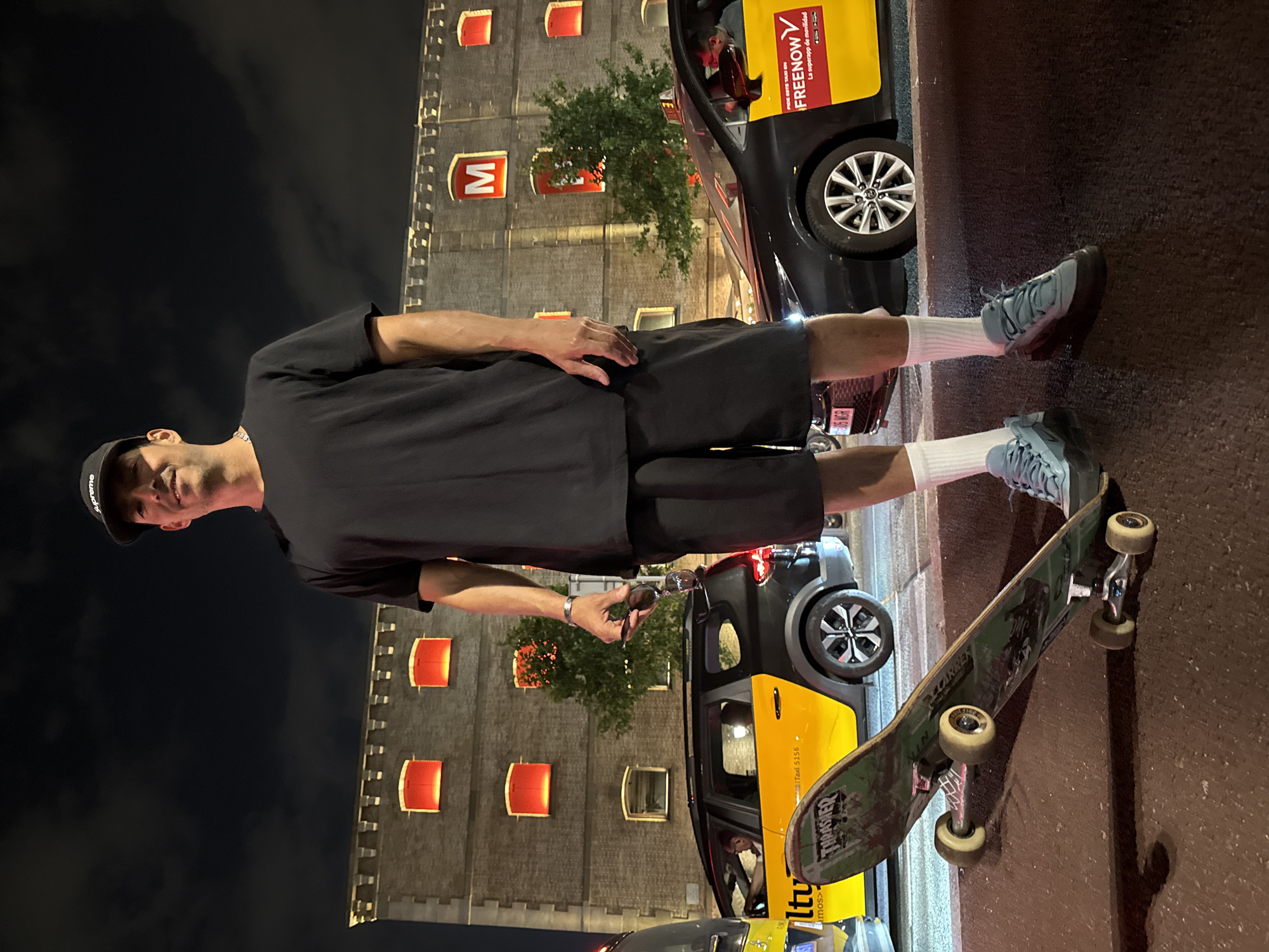 A man in dark shorts and a black t-shirt poses with a skateboard and sunglasses outside the Hotel Barceló Raval in Barcelona, Spain, at night. He wears white socks, light blue shoes, and a black hat. Two yellow and black "FreeNow" taxis are parked adjacent to the man. The hotel's facade, with its signature "M" logo and illuminated rectangular windows, is visible in the background against a dark sky. The scene is illuminated by ambient light.